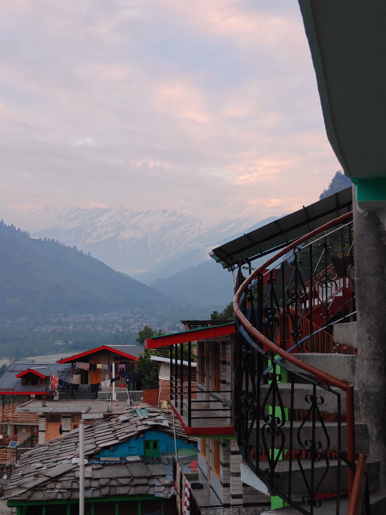 This media file shows a view of Vashisht, India, with a focus on the architecture and the surrounding mountainous landscape. In the foreground, a spiral staircase with a dark metal railing and a wooden handrail leads upwards. The staircase is constructed of concrete steps. Adjacent to the staircase are buildings with red and green trim, and stacked stone and brick walls. These structures appear to be part of guesthouses or homes, with visible laundry hanging from clotheslines on one of the rooftops. The rooftops themselves are covered with slate tiles, and one building features a vibrant blue exterior with a green door.
In the middle ground, a cluster of houses forms a village nestled in a valley. The houses are primarily brick with red roofs, and the surrounding area is covered in lush green trees. Further in the background, majestic snow-capped mountains rise, their peaks shrouded in a light haze. The sky above is filled with soft, scattered clouds, illuminated by the warm hues of either sunrise or sunset, suggesting either early morning or late evening. The overall mood is tranquil and scenic, highlighting the natural beauty of the Himalayas and the unique architectural style of the region. There is no visible text in the image.
