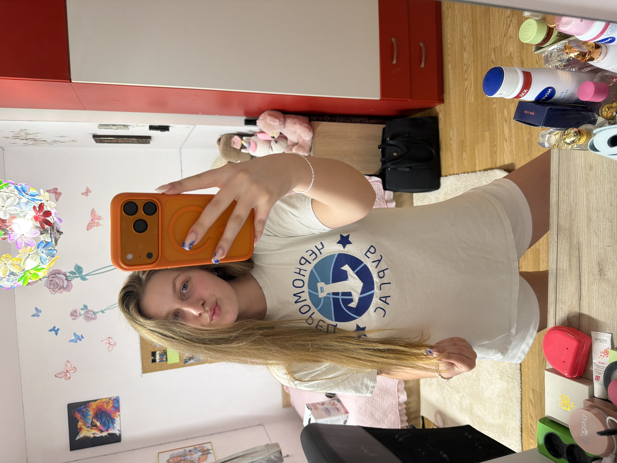 A young woman is captured in a mirror selfie indoors, likely in a bedroom or dressing area in Burgas, Bulgaria. She has long, straight blonde hair and is wearing a cream-colored t-shirt with a blue and white graphic, paired with white shorts. She holds an orange smartphone, obscuring part of her face, and her nails are painted with blue tips. A white beaded bracelet adorns her right wrist.
The t-shirt's graphic displays a circular logo with a stylized figure and the Cyrillic text "ПЕРФОРМАНС РЪГБИ" (PERFORMANCE RUGBY). The room reflected in the mirror reveals red and white wardrobe cabinets and light wooden flooring. To the left, a colorful light fixture resembling butterflies and flowers hangs on the wall, beneath which are wall stickers of pink roses and various butterflies. A framed picture of a colorful cat and a cork board are also visible on the wall.
On the right side of the mirror, a collection of personal care products and cosmetics is arranged on a surface. These include a white Nivea bottle with a blue cap, several perfume bottles, a small blue box possibly labeled "OLD SPICE", a red heart-shaped container, and products with visible labels such as "LXAM" and "posiblle". A green power strip is also present. On the floor, a black handbag sits near a white fluffy rug, and stuffed animals, including a pink teddy bear, are nestled by the wardrobe. The overall scene suggests a personal and organized living space.