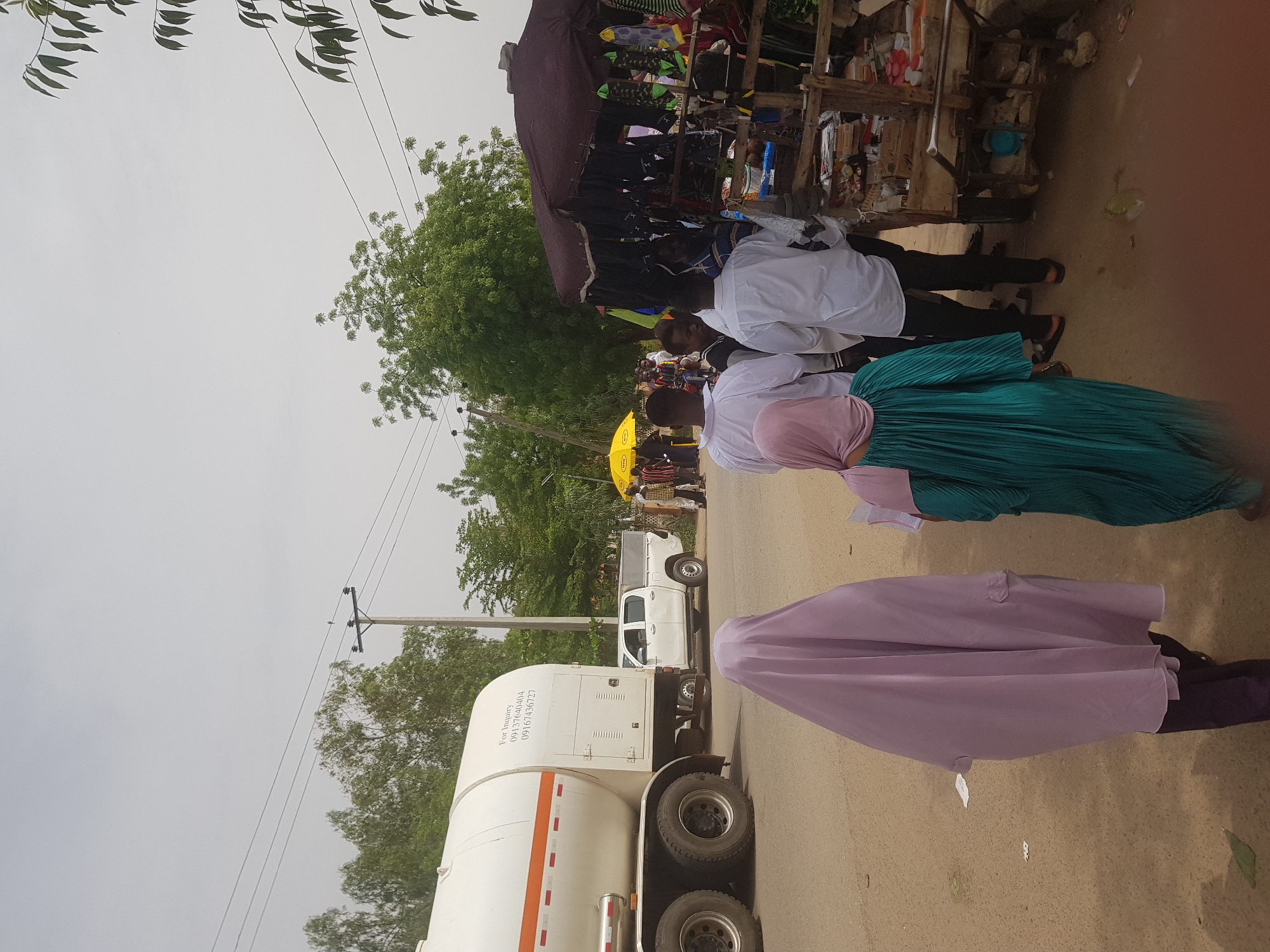 In the city of Dabawa, Nigeria, a tanker truck is parked on a paved road. Several people are present near a makeshift roadside stand. A woman wearing a pink hijab and a lavender outer garment is walking on the road. Another woman in a turquoise dress and pink hijab is standing near people wearing white shirts and black pants. A yellow umbrella is also visible near the people in white. The stand appears to be selling clothing and other items. Trees and power lines are in the background.
