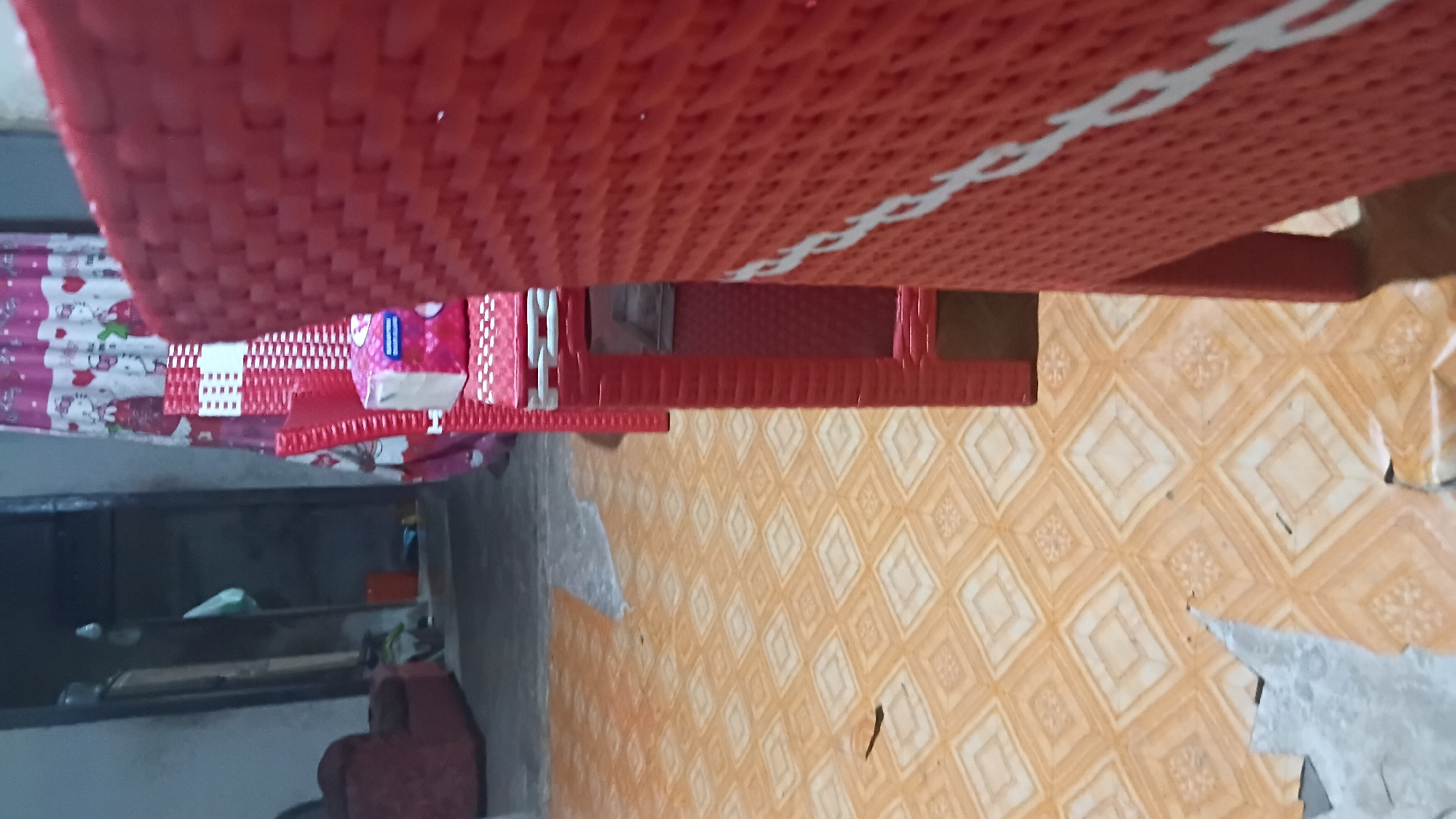 The image depicts an indoor scene with a tiled floor and red plastic furniture. The floor is covered in a pattern of light orange and beige squares with smaller decorative designs within them. To the right, a large red chair or stool made of woven plastic is prominently featured, filling a significant portion of the frame. In the background, a pink and white curtain with cartoon character prints hangs, possibly indicating a room within a home. A box of tissues is visible on a red chair in the middle ground. The environment appears to be a residential interior. The provided information states the location is Prabumulih, Indonesia.