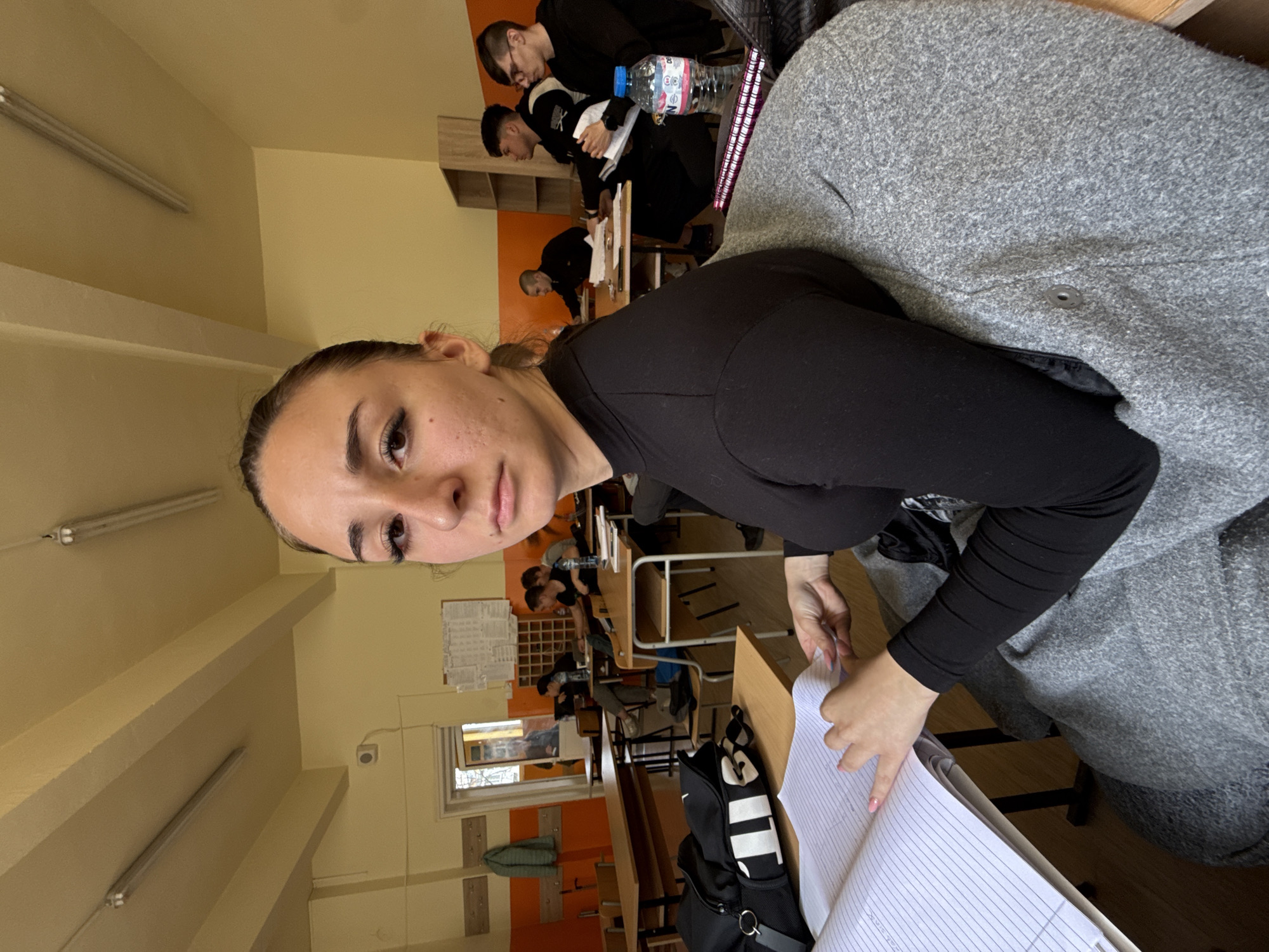 A medium-angle shot captures a young woman seated at a desk in what appears to be a classroom or lecture hall, possibly located in Sofia, Bulgaria, based on the architectural style and interior design typical of educational institutions in the region. She has fair skin, dark hair pulled back from her face, and is wearing a black long-sleeved shirt. The woman is looking directly at the camera with a neutral expression.
In front of her, on the desk, is an open notebook filled with lined paper, and a black bag with white accents. Her hands are visible, manipulating the pages of the notebook.
Behind her, several rows of desks are occupied by other students, most of whom are facing forward and appear to be engaged in writing or reading. Some are wearing dark clothing. A bottle of water is visible on one of the desks in the background.
The room is illuminated by fluorescent lighting, and the walls are painted in light yellow and orange hues. A window and a bulletin board are visible in the background. The scene suggests an academic environment, possibly during a class or examination.
