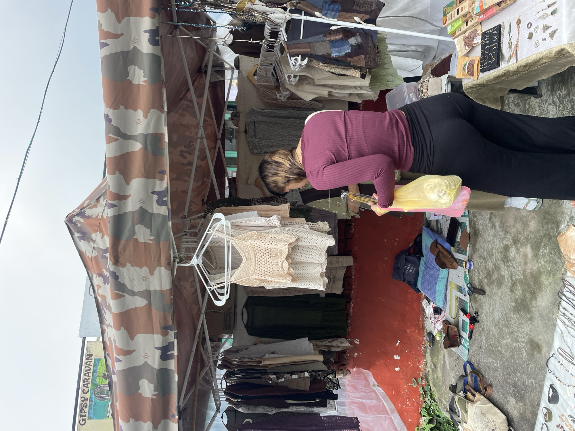 In Bhagsu, India, a woman with brown hair is browsing a stall displaying clothing and accessories. She is wearing a long-sleeved maroon top and black pants, holding a yellow plastic bag. The stall features a camouflage-patterned canopy, with various garments hanging on racks. Below the canopy, a red wall serves as a backdrop for more items displayed on the ground, including blankets, bags, and tools. To the right, a table is covered with jewelry and packaged goods. The scene suggests an outdoor market or fair.
