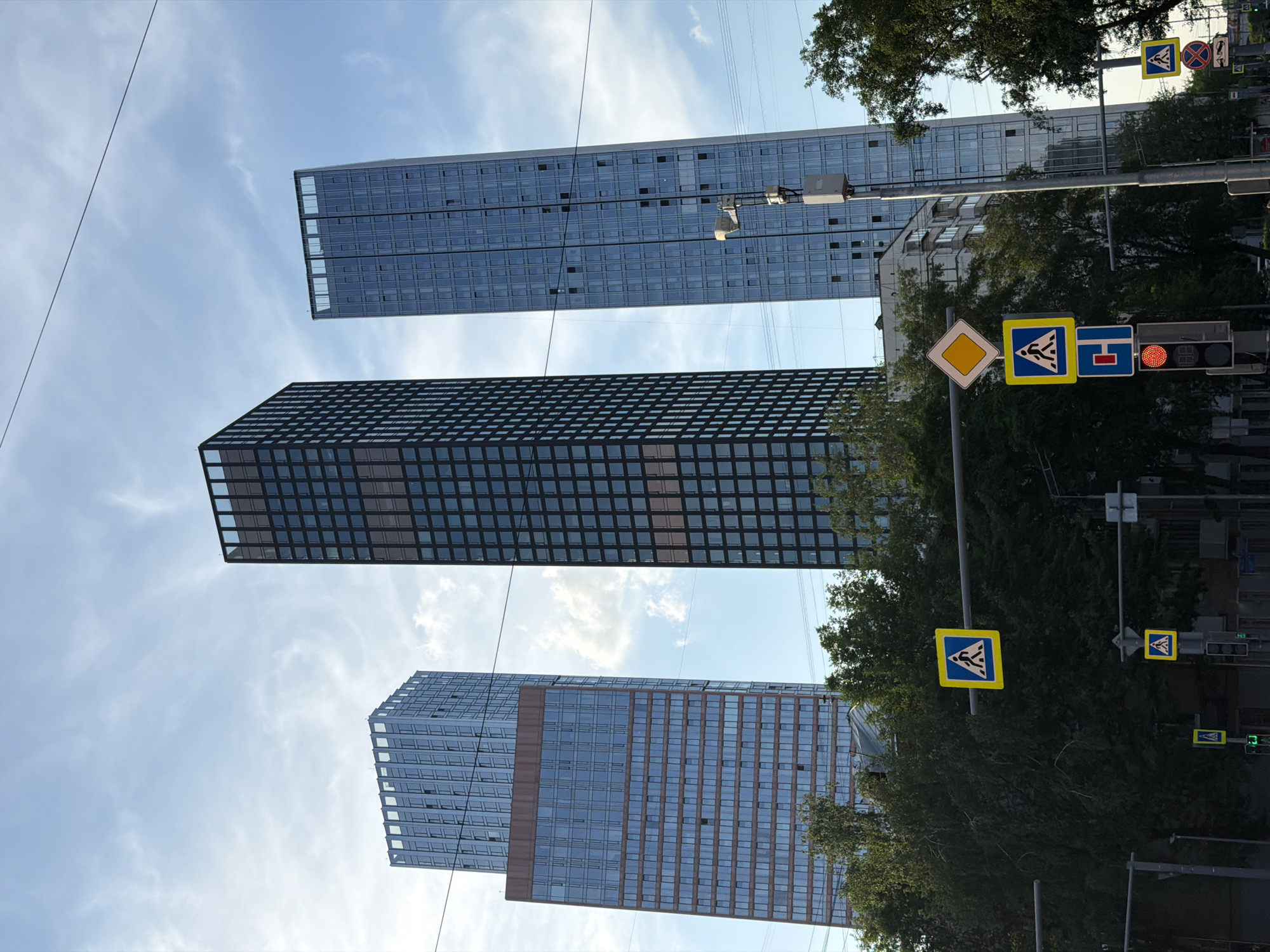 The image shows a street scene in Moscow, Russia, with a view of three modern skyscrapers. The buildings, constructed with glass facades, dominate the skyline. Street signs and traffic lights are visible in the foreground, along with trees. The traffic light is red.