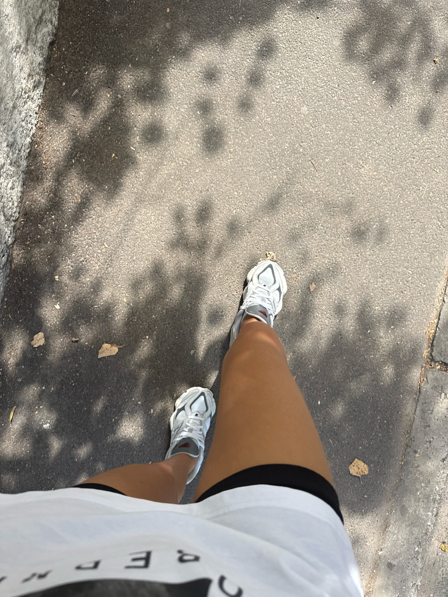 The image shows a low-angle perspective of a person's legs and feet walking on a paved surface, likely a sidewalk or path. The person is wearing white athletic shoes with grey accents and black shorts. A portion of a white t-shirt with black lettering is visible at the bottom of the frame. The ground is dark asphalt, with shadows of tree leaves dappled across it. There are a few fallen leaves on the ground. To the left, a textured, grey concrete wall runs along the edge of the path. The scene is outdoors, suggesting a park or urban street setting in Chișinău, Moldova, characterized by its paved walkways and surrounding vegetation.