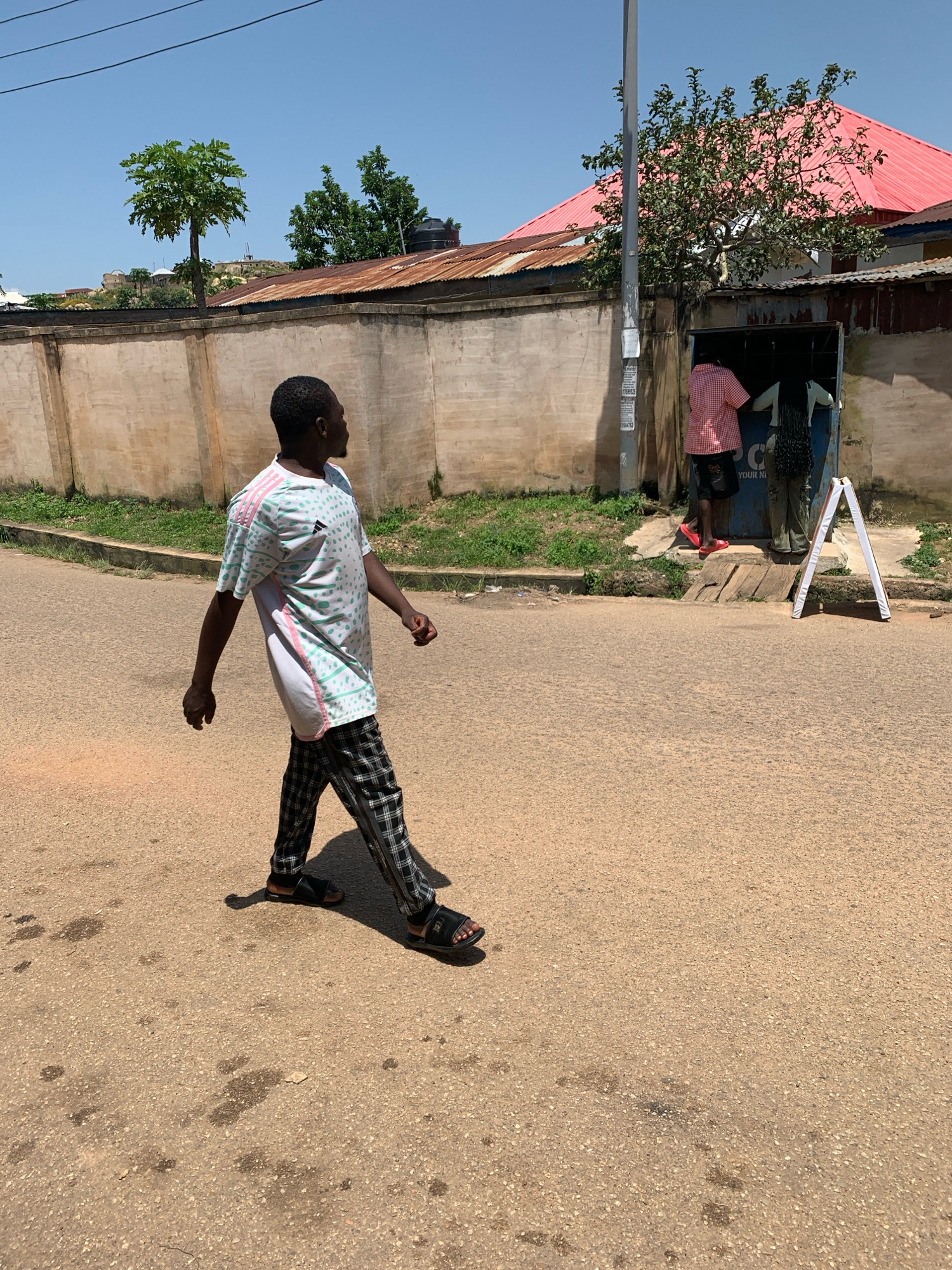 The image captures a street scene in Jos, Nigeria, on a bright, sunny day. A young man, dressed in a white t-shirt with a green and pink pattern and black and white checkered pants, walks from left to right across a paved road. He is wearing black sandals. To the right of the road, a small shop with a blue facade and a red corrugated metal roof is visible. Two individuals are partially visible inside the shop. One, wearing a red and white checkered shirt and dark shorts, stands near the entrance, while another, wearing olive-green pants, is further inside. A white A-frame sign stands on the sidewalk in front of the shop. A light-colored concrete wall runs along the background, topped with corrugated metal. Trees, including one with large leaves, grow behind the wall. The sky is clear and blue. The ground of the street shows several dark stains, possibly from oil or water.