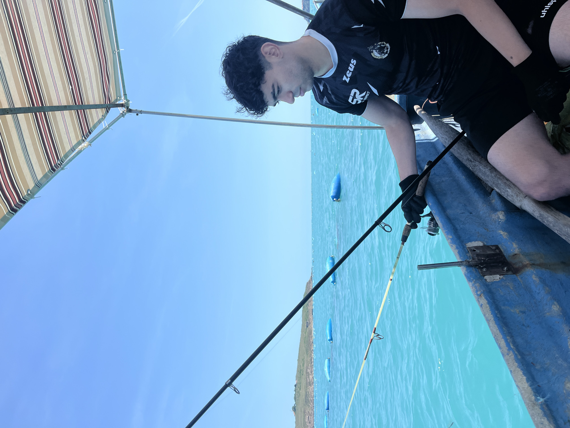 A young man is depicted from the torso up, fishing from a small, worn blue boat under a striped canopy on a clear, sunny day in an unknown aquatic location. The man, with dark, curly hair, wears a black short-sleeved athletic shirt with white accents and "SR" and "Zeus" logos, black shorts, and black gloves. He is focused on a dark fishing rod held in his hands, looking down and to his right. Another lighter-colored fishing rod is visible resting near the boat's edge. The water is a vivid turquoise-blue with small ripples. In the mid-ground, several large, blue cylindrical buoys float, with a low-lying, vegetated landmass and a distant small structure visible on the horizon to the left. The sky is a bright, cloudless blue with a faint contrail in the upper right. The boat shows signs of wear, with peeling paint and rust stains.
