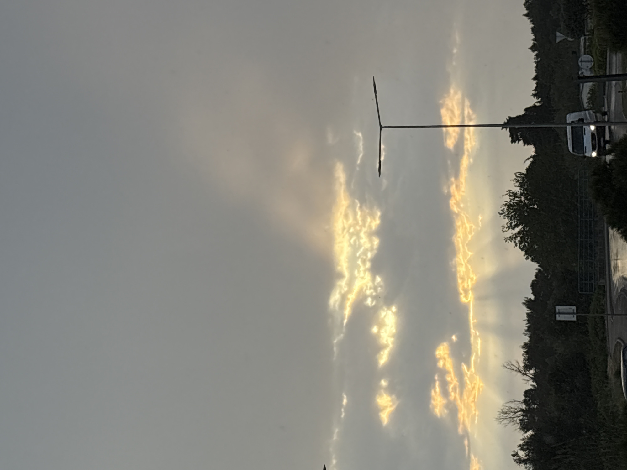 The image captures an outdoor scene, likely at dawn or dusk, in Bar, Montenegro. The sky dominates the upper and central portions, characterized by a mix of grey, overcast clouds and striking patches of golden sunlight. These sunlit clouds appear to glow brightly, creating dynamic streaks of light that suggest either crepuscular rays or direct sunlight breaking through a dense cloud cover during the golden hour.
A tall, dark utility pole or street lamp with a horizontal crossbar stands vertically in the mid-ground, silhouetted against the bright sky. Below the level of the glowing clouds, a dense line of dark green trees and foliage stretches across the lower part of the scene, indicating a natural or landscaped boundary alongside a road. Partially obscured by the foliage, a white van is visible on what appears to be a paved road, with its headlights illuminated, further emphasizing the low light conditions of early morning or late evening.
The overall impression is one of dramatic natural light, highlighting the contrast between the dark elements of the landscape and the vivid, glowing sky. There is no discernible text visible in the image.
