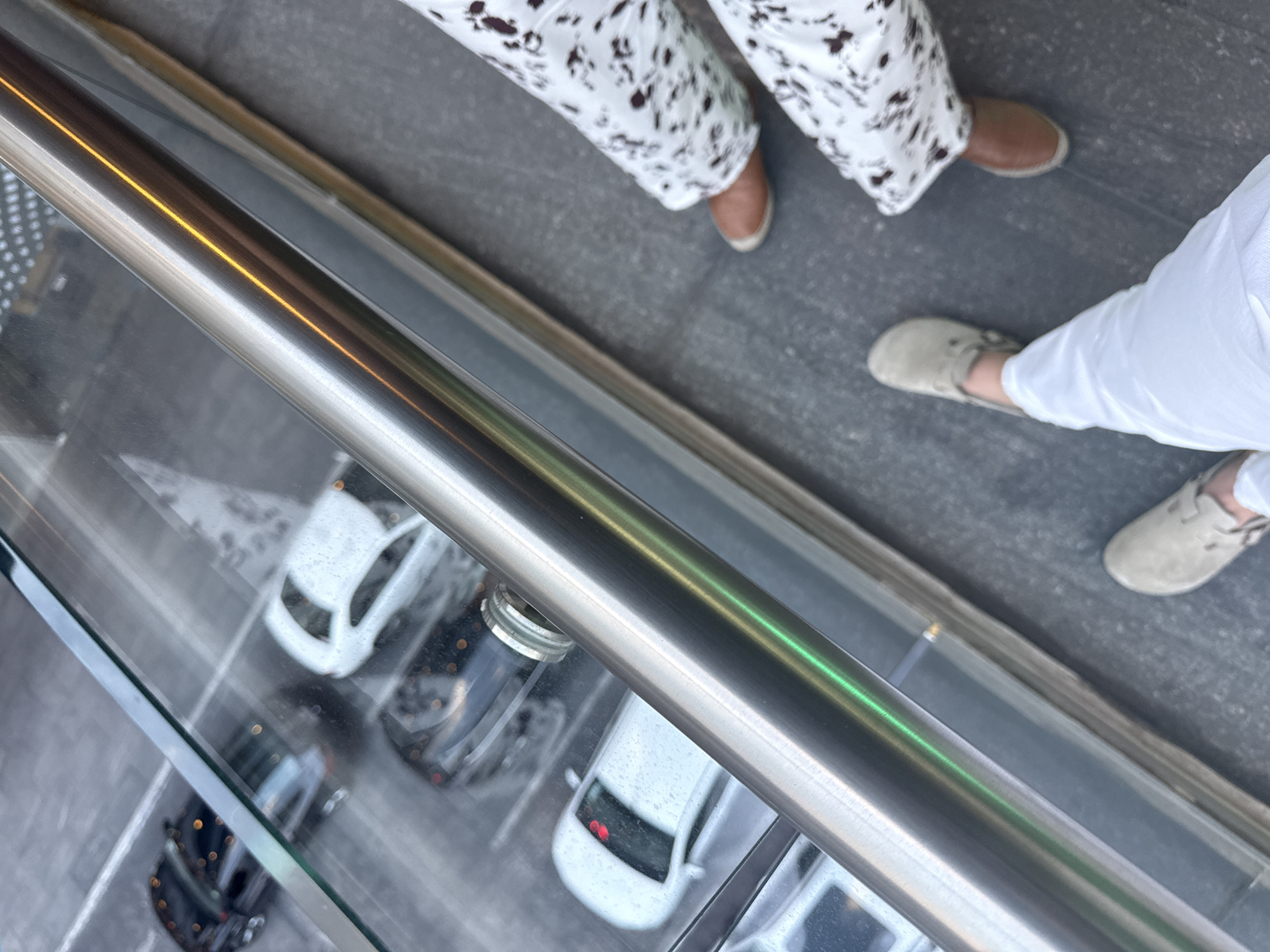 This image, taken from an elevated perspective, shows the lower half of two individuals standing on a paved surface, likely an outdoor walkway or balcony, next to a clear glass barrier with a metal railing. Below the glass barrier, several cars are visible, parked in what appears to be a parking lot or street.
The individual on the left wears white pants with an irregular dark pattern and brown, low-cut shoes. The individual on the right wears plain white pants and light beige or grey casual shoes. Both appear to be standing still or walking slowly.
The metal railing, positioned prominently in the foreground, reflects light with streaks of warm yellow/orange and cool green hues. The surface the people are standing on is a dark grey, possibly asphalt or concrete. Through the transparent glass panel below the railing, several vehicles, including white and dark-colored cars, are distinctly seen parked on a grey, slightly textured ground.
The lighting suggests it is daytime. No specific weather conditions are clearly discernible, but the overall illumination is bright. The scene depicts a common urban or suburban setting, consistent with the provided location context of San Bernardino Tlaxcalancingo, Mexico, showing pedestrians observing or passing by a scene below. No text is visible in the image.