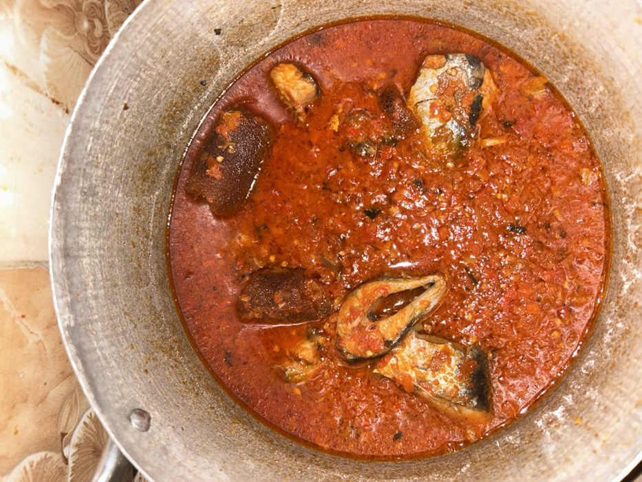 A top-down shot features a metal pot filled with a vibrant red stew. The stew contains large chunks of fish and what appear to be pieces of animal skin or meat, possibly cow feet. The texture of the sauce is chunky and somewhat oily. The pot's interior shows wear and discoloration, suggesting it has been used frequently. The surrounding surface is tile and appears to be well-worn.