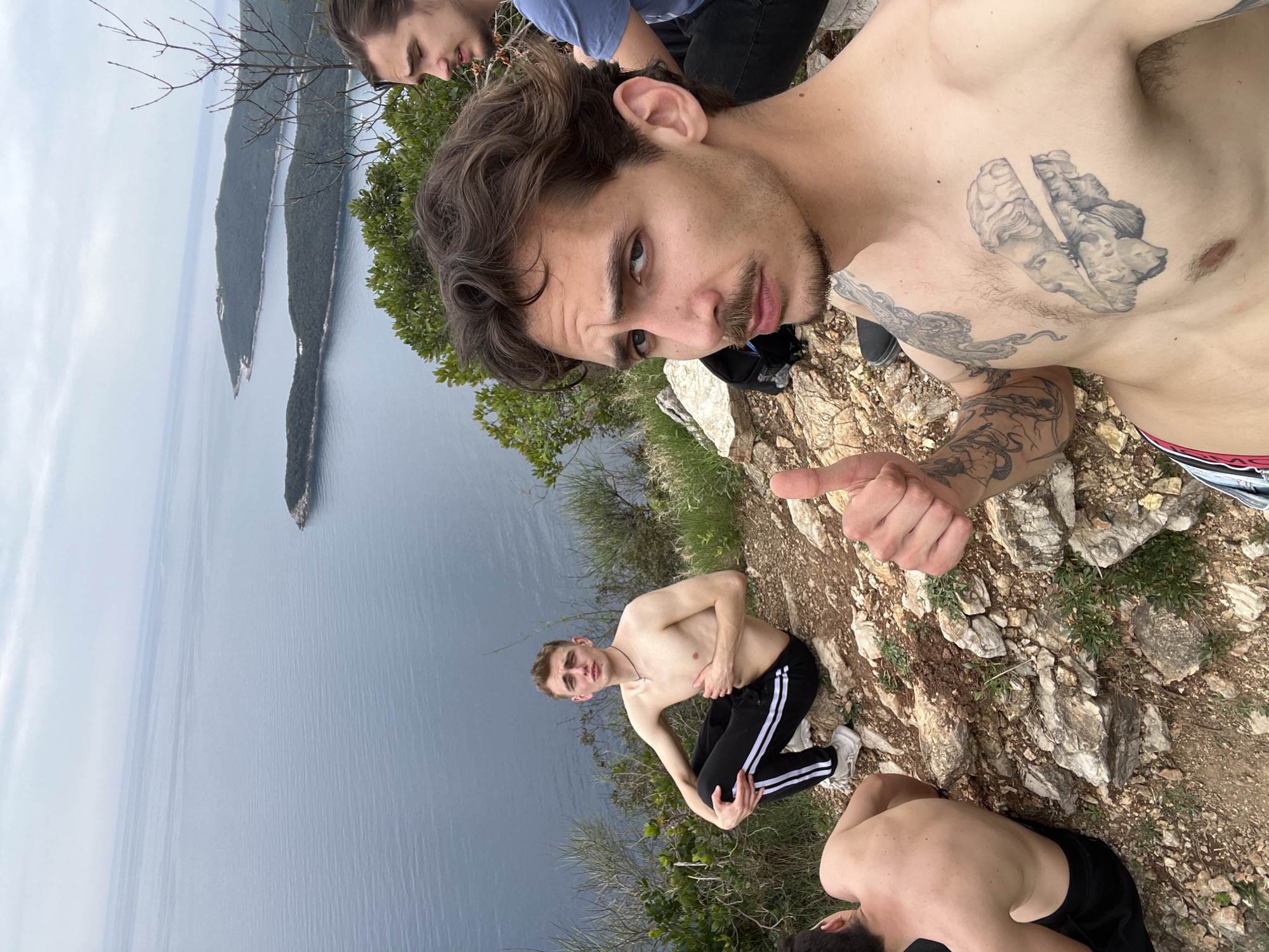 A selfie-style photograph shows four people on a rocky precipice with a view of the Adriatic Sea and land formations, likely taken in the vicinity of Sveti Stefan, Montenegro. One man, shirtless with tattoos on his chest and arms, is holding the camera and giving a thumbs-up. To the left and slightly behind him, another person is partially visible, wearing a blue shirt. Below the first man, a second shirtless individual is kneeling, looking up at the camera. Part of a fourth person's torso is visible at the bottom of the frame. The background features a body of water and landmasses jutting into the sea.