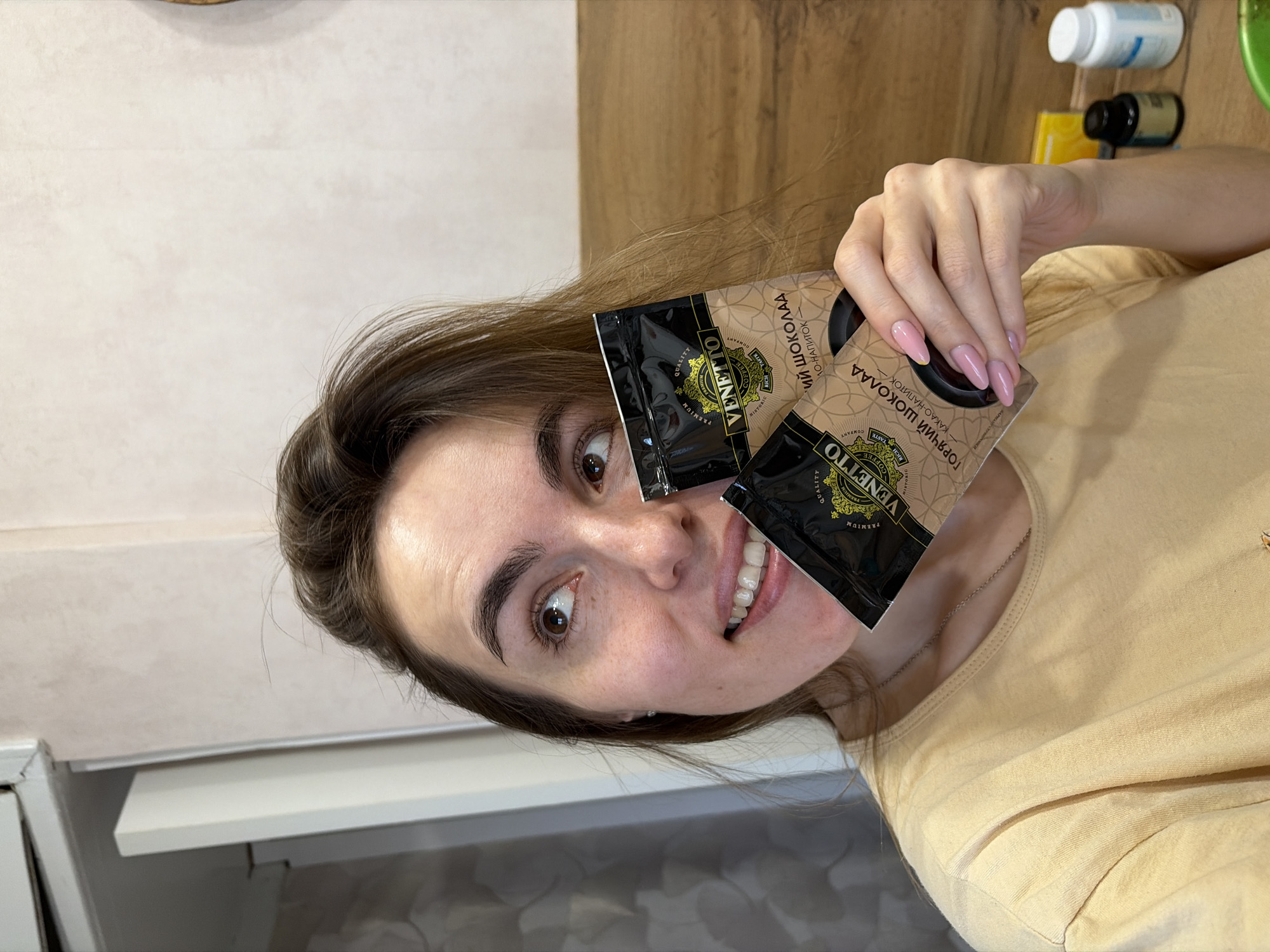 A woman in a light yellow T-shirt is holding two black packets of "VENETTO" hot chocolate mix close to her face, partially obscuring her left eye and cheek. The packets are labeled in Cyrillic script and English. The woman has fair skin, brown hair, and light pink nail polish. She is smiling, and her eyes are looking towards the camera. The background consists of a kitchen counter with light-colored cabinets and a light wooden surface. Several bottles and a yellow box are visible in the upper right corner of the frame.