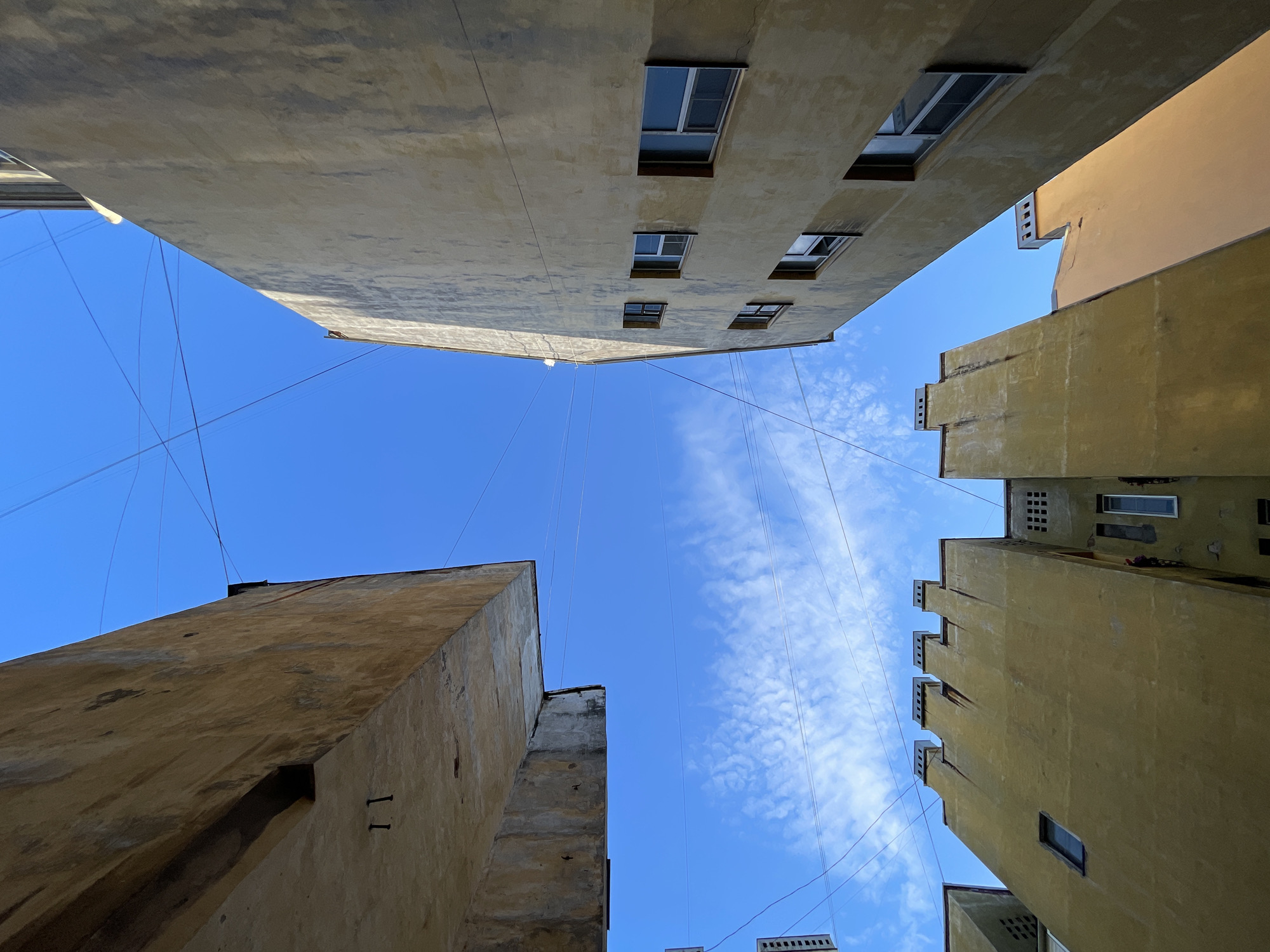 The image captures an upward perspective of buildings and the sky in Saint Petersburg, Russia. Several buildings, primarily with yellow-toned facades, are visible, their architectural styles suggesting pre-revolutionary or early Soviet construction. The buildings are densely packed, creating a sense of an urban courtyard or a narrow street.
In the foreground, the underside of a concrete structure, possibly a balcony or an overhang, is prominent. It has a rough, weathered texture with visible rebar or fasteners and the numeral "11" is crudely inscribed on its surface.
To the right, a tall building with a pale yellow facade rises, featuring several uniformly spaced windows. Some windows are open, revealing reflections of the sky. The facade shows signs of age and weathering, with streaks and discoloration.
In the lower center and left, another building with a similar yellow facade is depicted. Its roofline is distinct, with a series of stepped, almost crenellated, protrusions. Several windows are visible on this building as well, some small and square, others rectangular.
The sky above is a vibrant blue, with scattered, wispy white clouds. A complex network of overhead wires crisscrosses the sky, a common sight in older urban areas.
The overall impression is one of a specific urban environment, likely an older part of Saint Petersburg, characterized by its distinct architecture, close-set buildings, and the ever-present sky adorned with utility lines. There are no people visible in the frame.