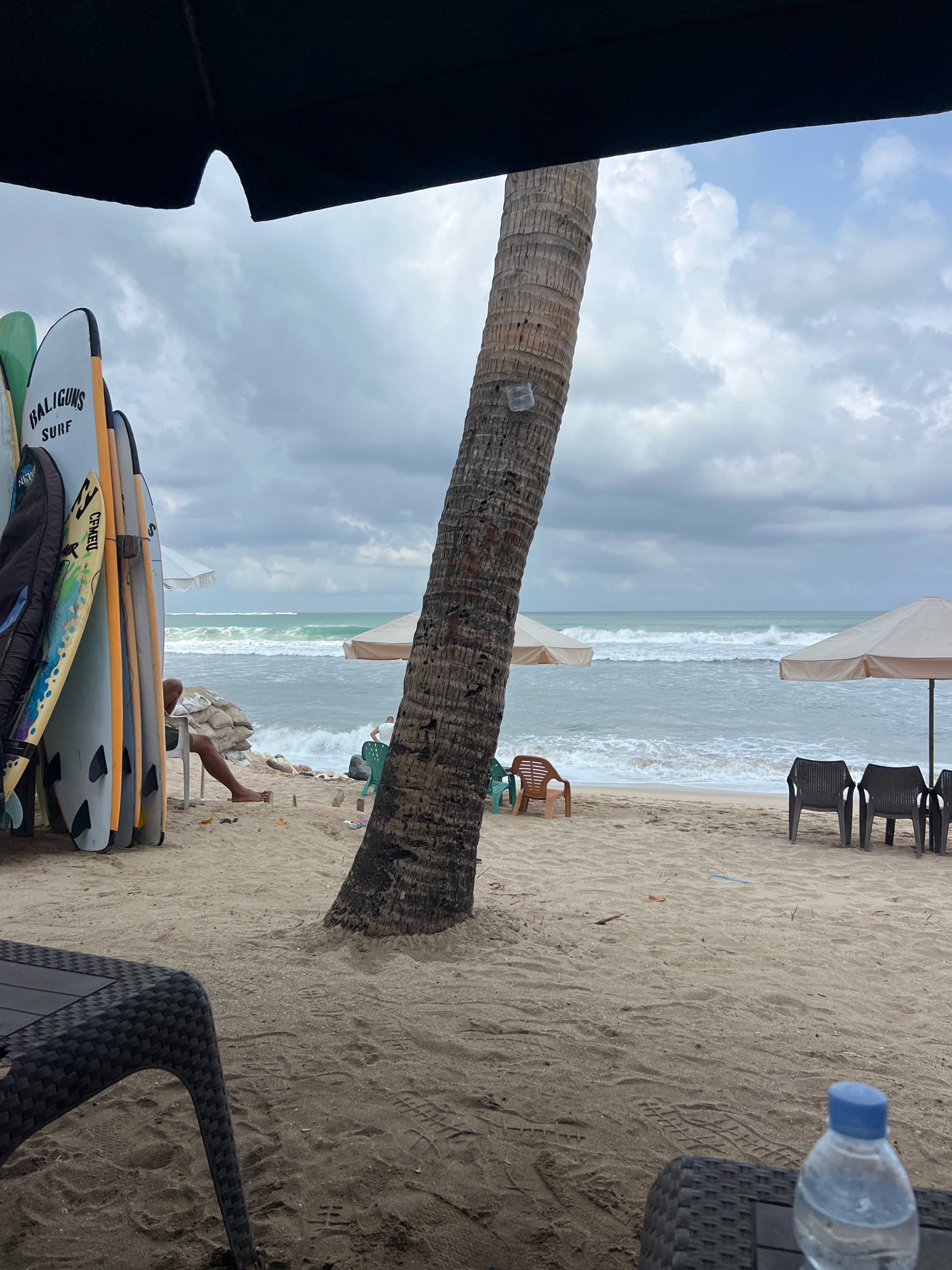 This image depicts a beach scene in Kuta, Indonesia, likely during overcast weather. In the foreground, a dark umbrella shades a collection of surfboards leaning against each other. The surfboards are of various colors and designs, with some bearing branding like "BALIGUNS SURF." Further into the scene, a large palm tree trunk is visible on the right. The sandy beach stretches towards the ocean, where waves are breaking under a cloudy sky. Several beach chairs and umbrellas are scattered on the sand. A person is seated on a white chair near the surfboards, appearing to rest. Another person is visible in the distance near a beach umbrella. In the immediate foreground, a portion of a black woven table is visible, with a plastic water bottle on it. The overall impression is a relaxed beach atmosphere, possibly a surf rental or beachside cafe area.