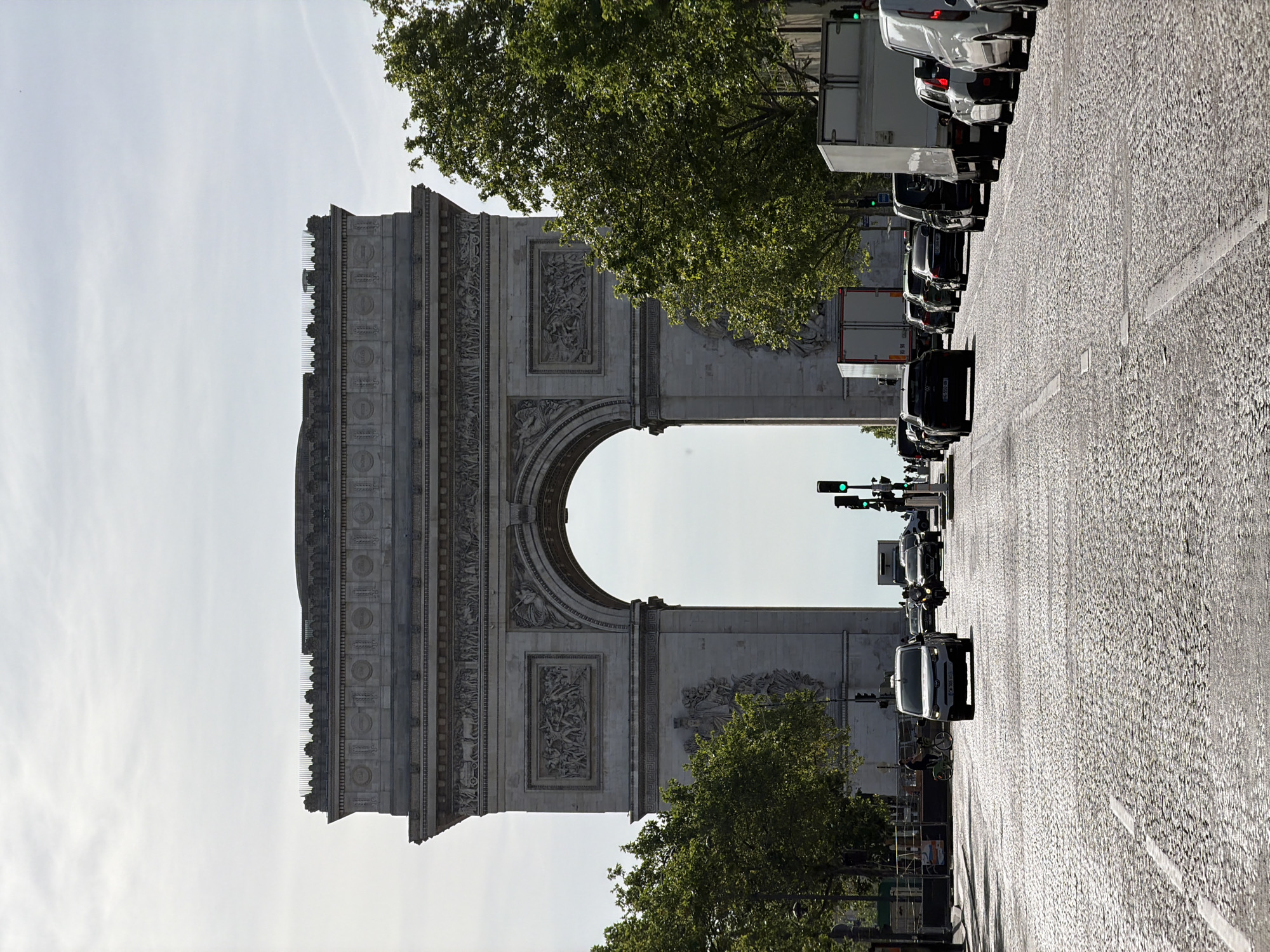 The image depicts the Arc de Triomphe in Paris, France. The perspective is from a street view looking directly at the monument. The street is lined with vehicles, and there are trees along the sides. The sky is visible through the arch. The traffic lights are green.