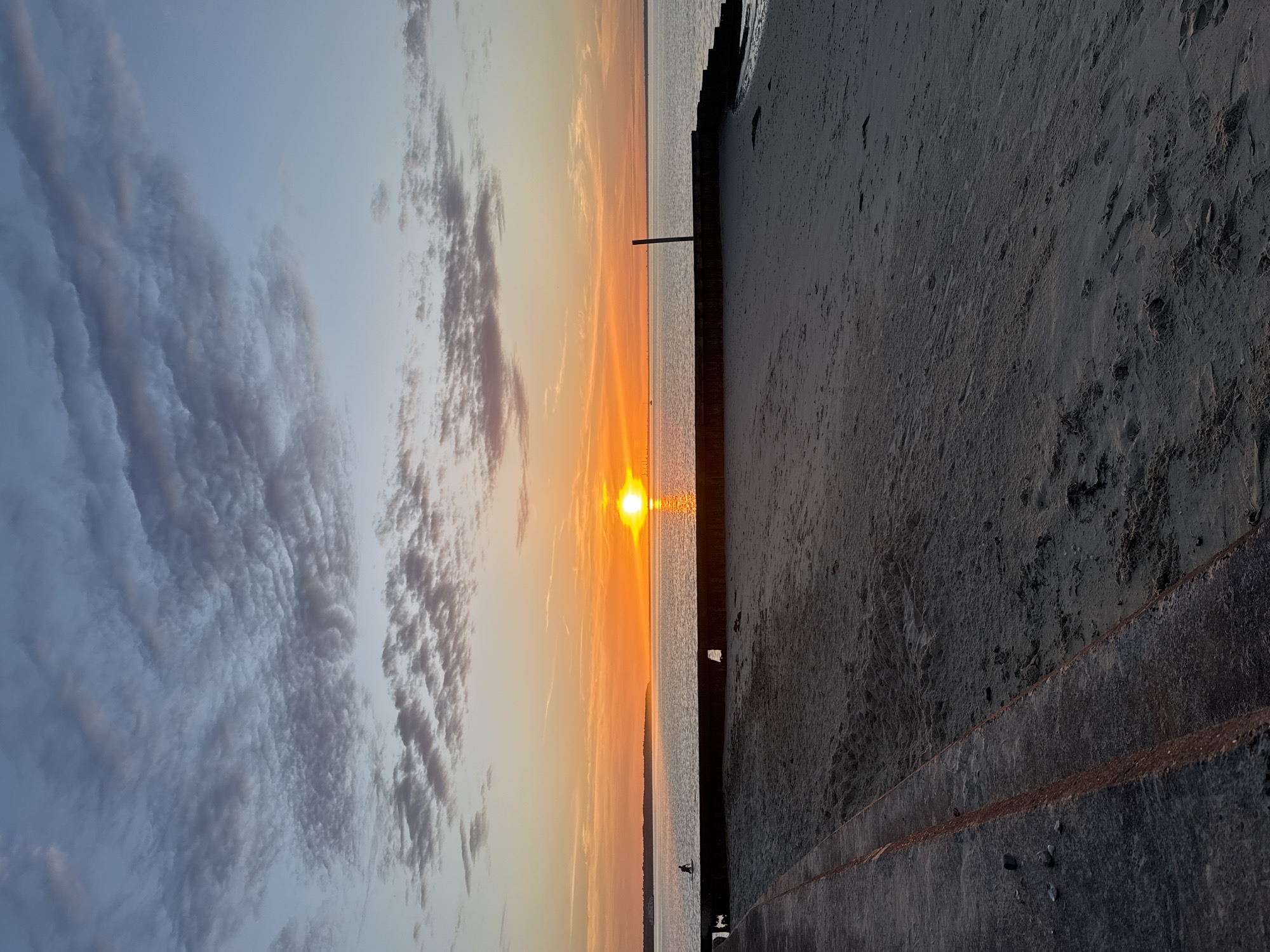 The image shows a sunset at Ryde, United Kingdom. The sun is setting over the sea, reflecting in the water. There are clouds in the sky, tinted with orange hues from the sunset. A concrete seawall or steps are visible on the right side of the frame, leading down to a sandy beach. A thin wooden post or pylon stands vertically in the water near the seawall. A person is standing in the sea near the shoreline.