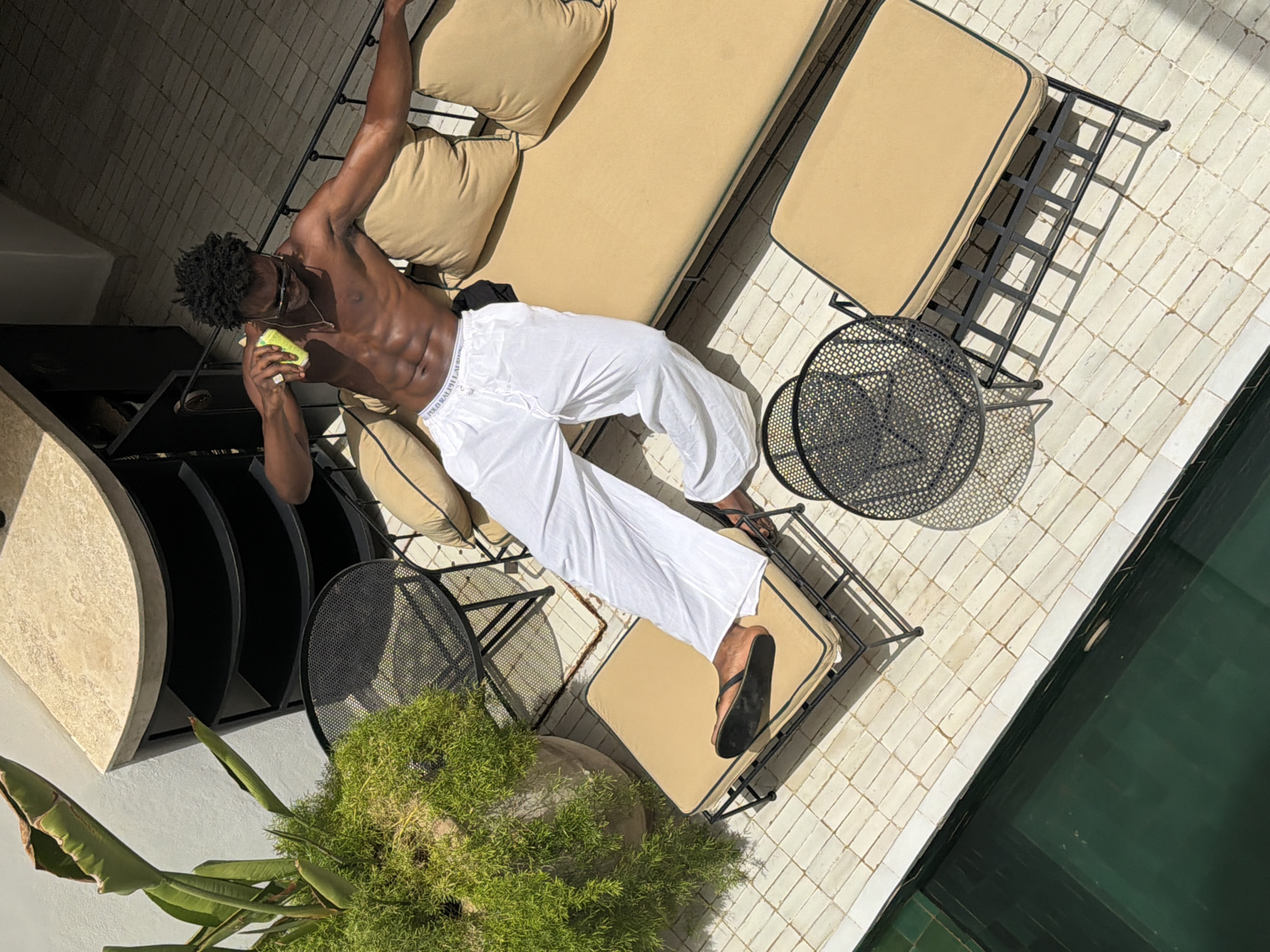 An overhead shot captures a shirtless Black man with muscular abs and dark, curly hair lounging on a tan outdoor sofa. He wears white lounge pants with a visible Polo Ralph Lauren waistband and black flip-flops. In his right hand, he holds a light-yellow beverage can. He is outdoors, with a pool to his left, framed by light-colored tiled flooring. To his right are two black metal mesh tables and a large potted plant with green foliage. The setting appears to be a rooftop terrace or patio in Marrakesh, Morocco, evident from the architecture and the style of the outdoor furniture and tiling.