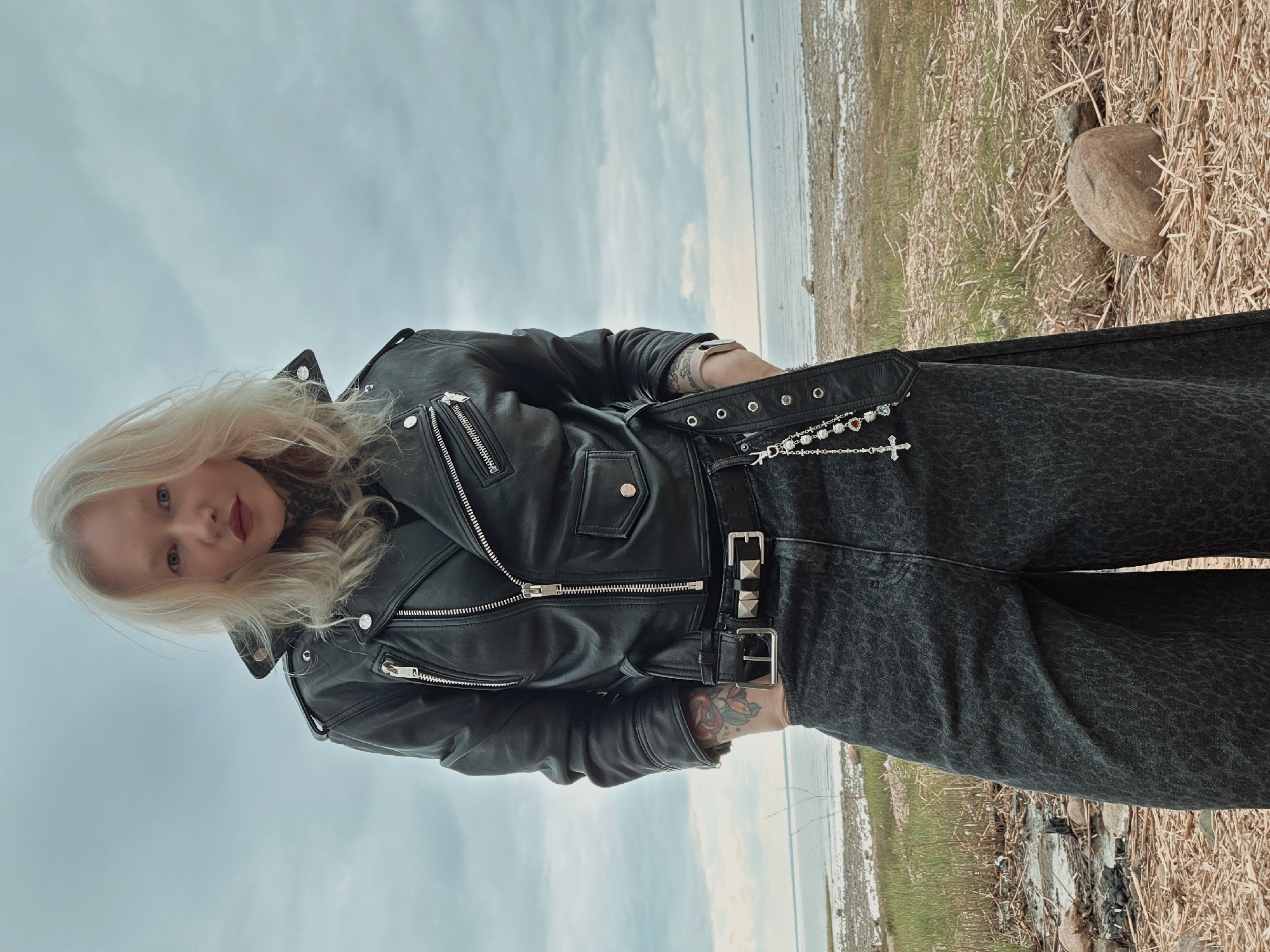 A waist-up portrait shows a pale-skinned person with blond hair standing on a grassy shore near a body of water. The person wears a black leather biker jacket over a dark-colored outfit, accessorized with a studded belt and a jewelry chain featuring a cross. Their tattooed arms are visible. The background features a cloudy sky, a body of water, and a shoreline with grass and small rocks. The exact location in the city of Unknown, Unknown, cannot be determined.