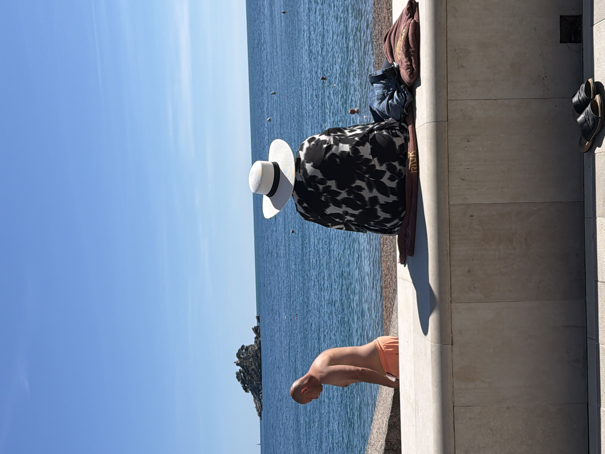 The image shows a scene at Petrovac centar, Montenegro, along the seaside. A man in orange swim trunks leans over a stone wall, presumably near the water. On the wall beside him are personal belongings including a woman's hat, a black and white patterned garment, a brown towel, a denim item, and shoes. The sea is visible behind him, with a small rocky island in the distance. The sky is clear and blue.