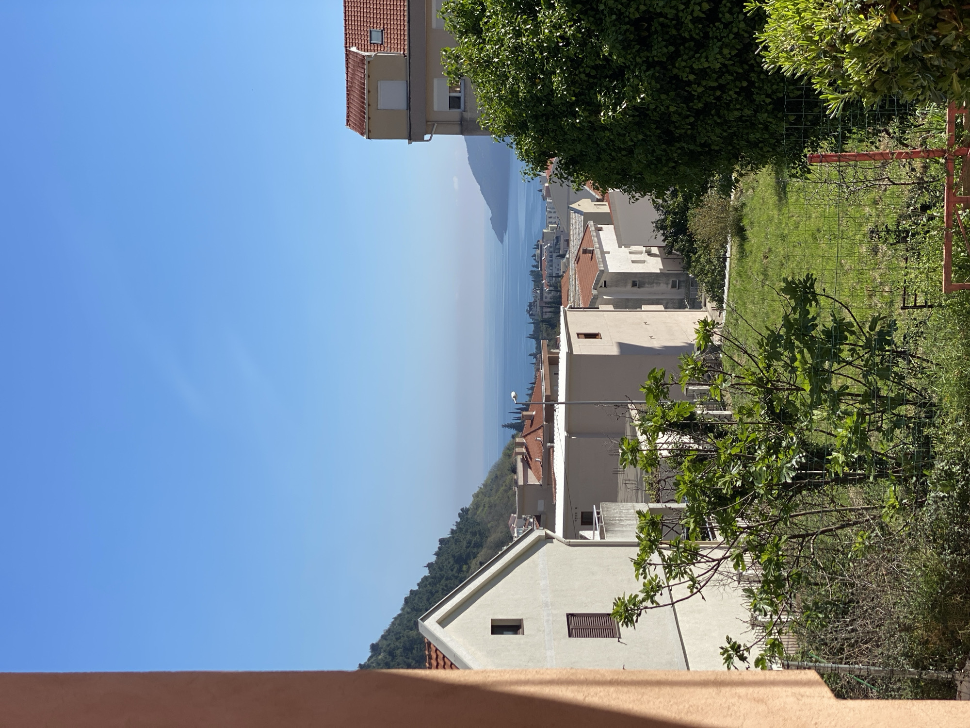 The scene depicts a view in Budva, Montenegro, oriented towards the Adriatic Sea. A residential area is seen on a sloping terrain, with buildings of various heights and architectural styles. The foreground features a glimpse of a building structure, possibly a wall or a roof, and shrubbery. Further into the scene are residential buildings and a green, grassy area enclosed by a fence. The sea stretches out to the horizon under a clear, blue sky. Haze obscures the distant shoreline. There are no people visible in the image.