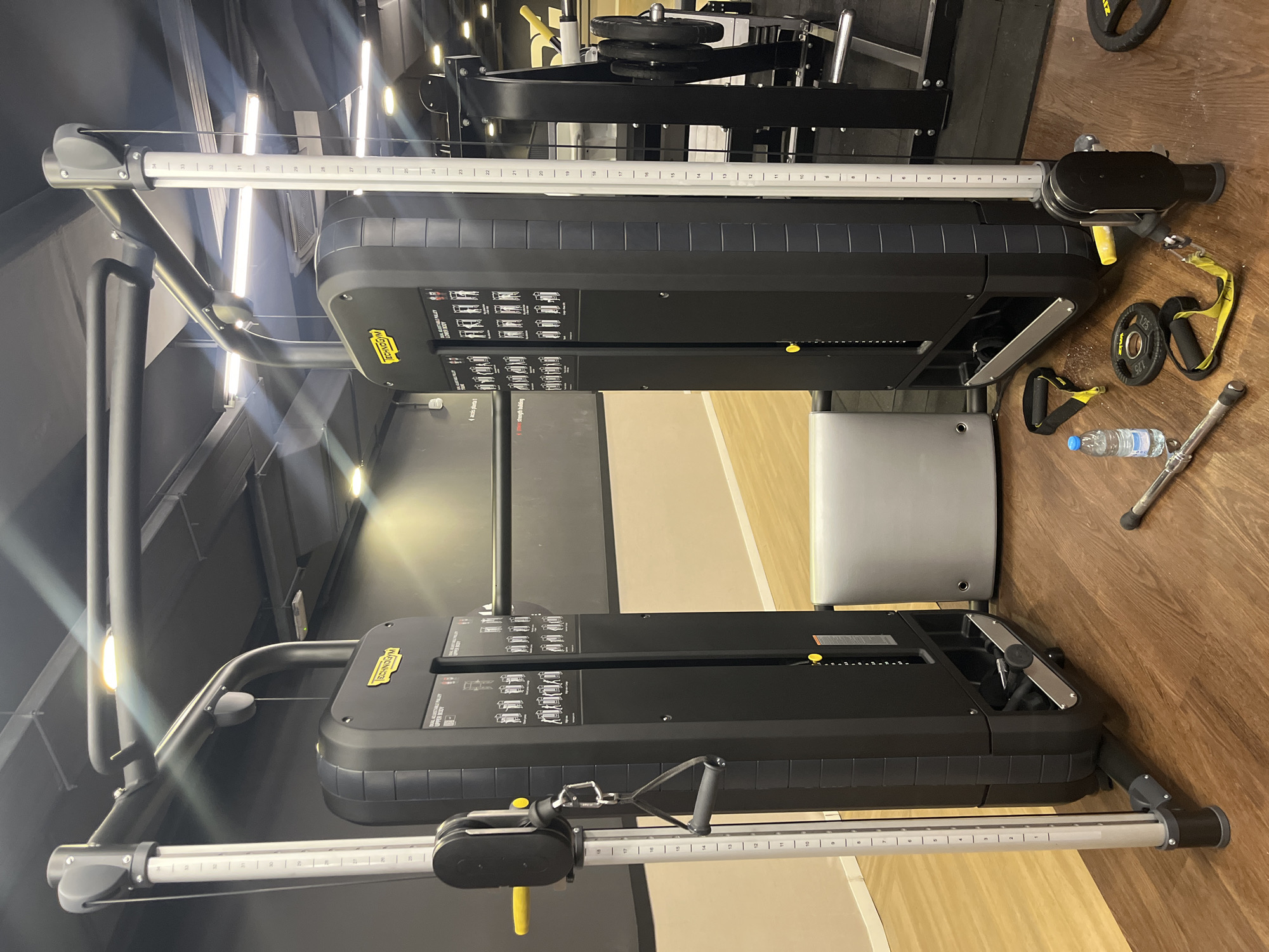 The image shows two Technogym dual adjustable pulley machines in what appears to be a gym setting. The machines are black with yellow accents and feature weight stacks and adjustable cable systems. The left machine has an attachment connected to the cable, while the right machine has several attachments and a 1.25 kg weight plate on the floor nearby, along with a water bottle. The flooring is wood-patterned, and the background is a combination of black walls and beige flooring. The environment suggests an indoor commercial gym. There are no indications in the image that confirm the location is Barcelona, Spain.