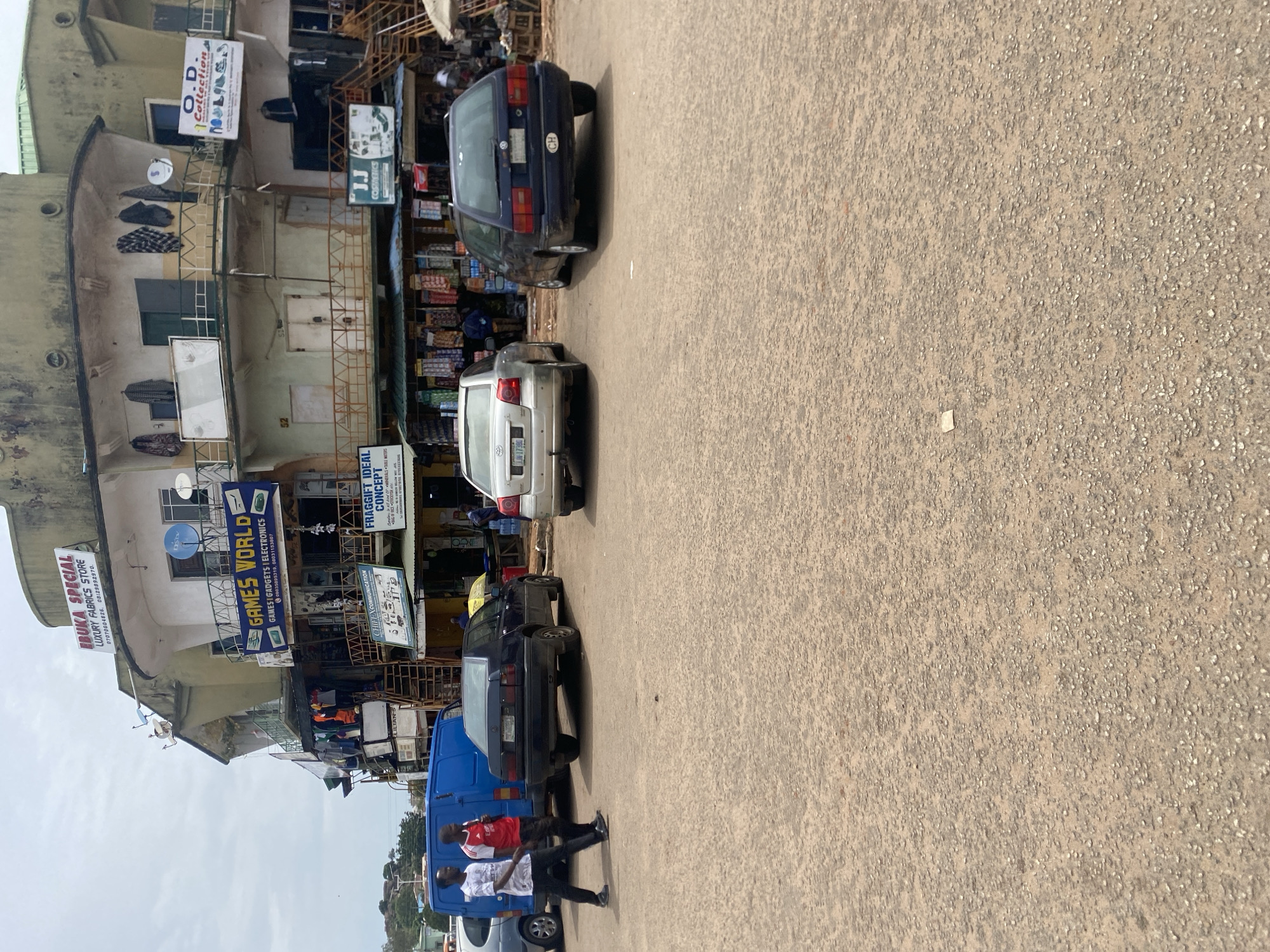 This is a medium-shot, eye-level, exterior photograph of a street scene in Jos, Nigeria, on a cloudy day. In the foreground, a dusty, unpaved road dominates the lower half of the frame. The midground features three cars parked on the road. From left to right, a dark blue van is partially visible, followed by a dark sedan. To the right of the sedan is a dark blue station wagon.
The background consists of a multi-story building that appears to be a commercial structure. The building has multiple levels with balconies and shops on the ground floor. Signs are visible on the building, including "EBUKA SPECIAL LUXURY FABRICS STORE," "GAMES WORLD GAMES | GADGETS | ELECTRONICS," "CHIEF X COMMUNICATION," "FRAGIGIFT IDEAL CONCEPT," and "J.J COSMETICS." Clothing and various goods are displayed outside some of the shops. Two men, dressed in casual attire, are walking on the road to the left of the frame, heading towards the building. The overall scene depicts a typical street environment with commercial activity and vehicles in Jos.