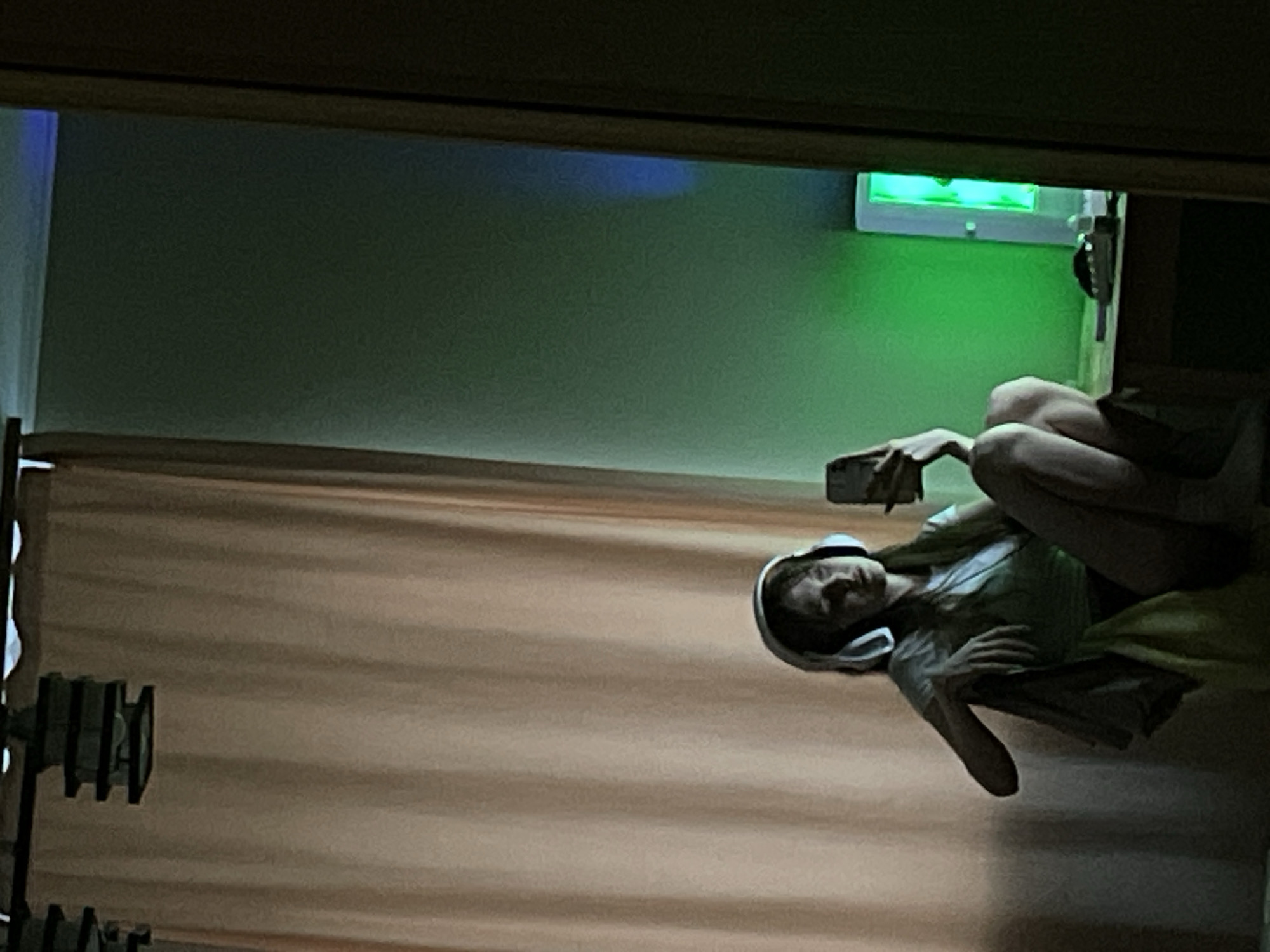 A person wearing headphones is seen holding a cell phone in what appears to be an indoor setting. The environment consists of what looks like a wall with a green light fixture. A curtain or wall is visible in the foreground. There is no indication of the location being the city of Ufa, Russia, or any specific location information.