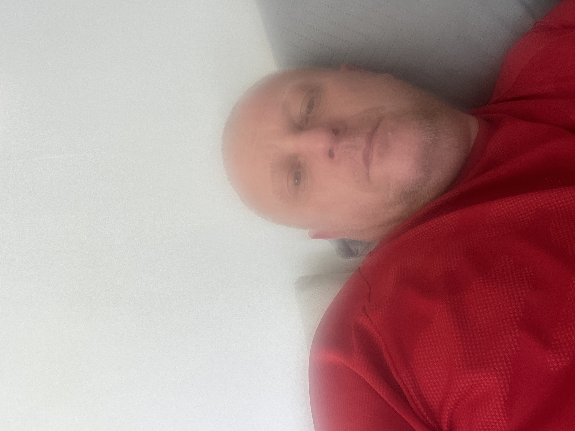 A Caucasian male with a shaved head and light stubble is lying on his back, wearing a red athletic shirt. He appears to be resting on a grey pillow or cushion. The background is predominantly white and slightly blurred. The exact location within Basingstoke and Deane, United Kingdom cannot be determined from the image.