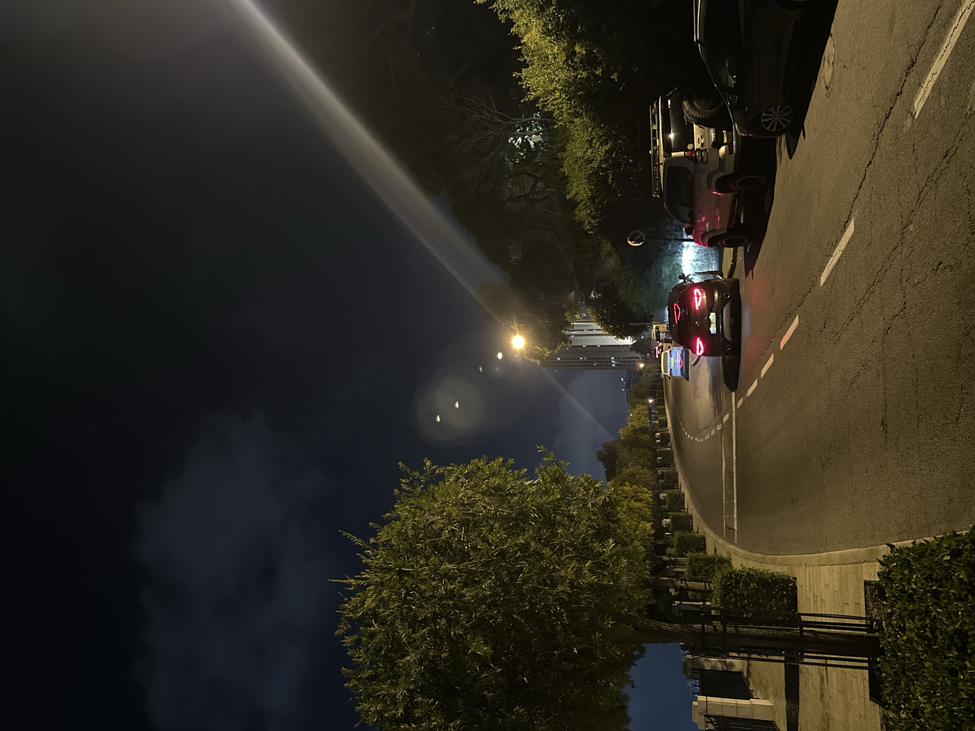 The image captures a nighttime scene on a curved road in Haifa, Israel. The road, illuminated by streetlights and vehicle headlights, is lined with trees and manicured bushes. Several cars are visible, including a dark SUV parked on the right side of the road, a light-colored SUV with a roof rack also parked on the right, and a dark-colored sedan driving away from the camera. Further up the road, another vehicle with illuminated taillights is also visible. The sky above is dark, with some clouds and a bright beam of light emanating from an unseen source, possibly a spotlight or a projector. In the background, a tall building can be seen. The overall atmosphere is urban and nocturnal.
