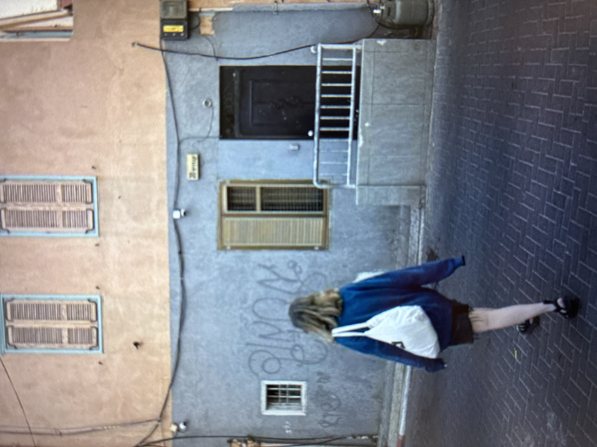 A full outdoor shot of a Bat Yam, Israel, street. On the left side is a peach-colored building with two window with blue frames. To the right is a taller blue building with two windows and a black front door. On the brick sidewalk is a woman wearing a blue sweatshirt, black shorts, white bag and sandals, walking down the sidewalk.