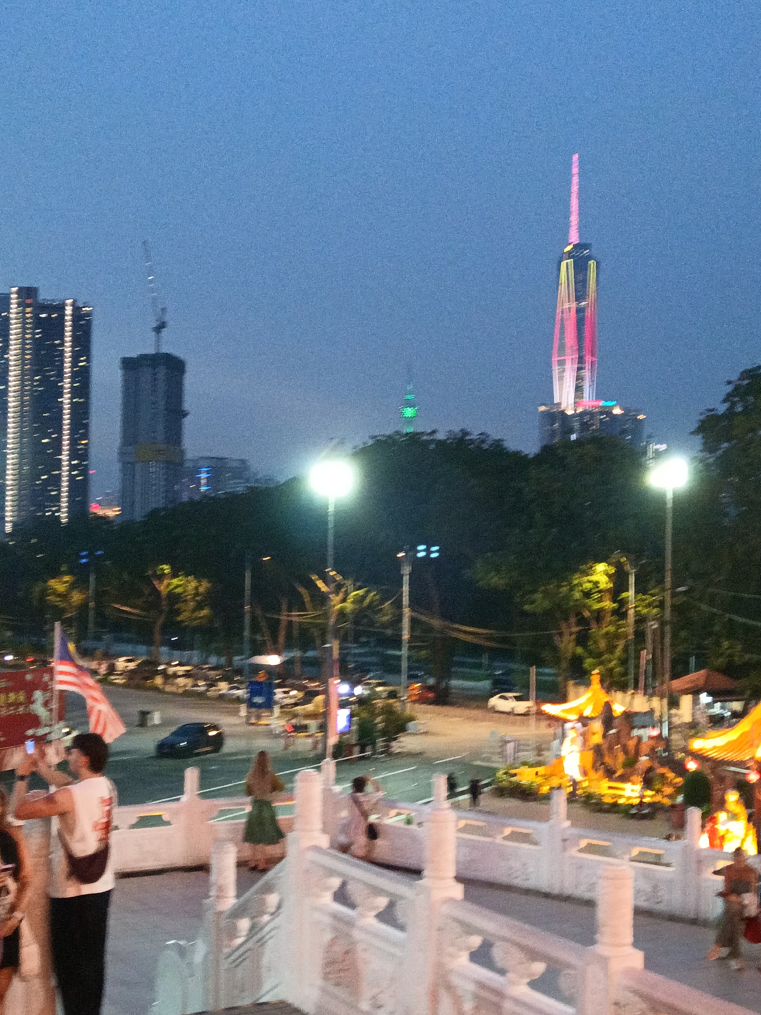 This is a medium-distance, eye-level shot of a city skyline at dusk, likely in Kuala Lumpur, Malaysia. In the background, the illuminated Menara Kuala Lumpur, also known as the KL Tower, glows green, and further to the right, the Petronas Twin Towers are lit with pink and yellow lights, signifying a vibrant city atmosphere. In the foreground, there is a white railing and steps, suggesting the vantage point is from an elevated area, possibly a park or observation deck. Several people are visible, including a man in a white shirt holding a phone and taking a photo. A Malaysian flag is prominently displayed on a pole. The lighting indicates it is twilight, with streetlights and building lights illuminating the scene. The environment appears to be a public space with trees and traffic visible on a road.
