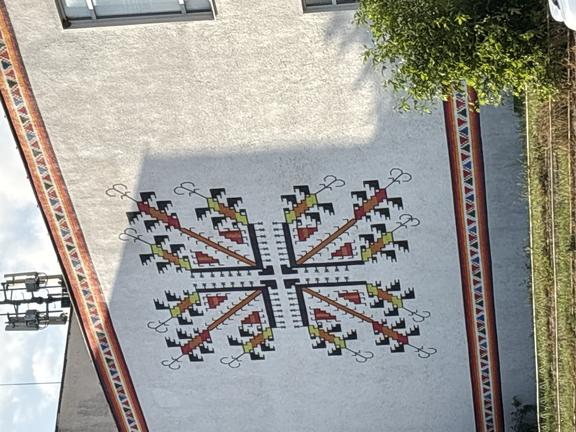 A low-angle, sunlit shot depicts a building facade in Garmen, Bulgaria. The facade is adorned with a large, symmetrical, cross-shaped geometric design featuring black, yellow, orange, and red elements. A colorful, triangular-patterned border trims the building's edges. A window is visible at the top of the frame, and foliage is present on the right side. Power lines and a utility pole are visible on the left. The building's surface is textured.