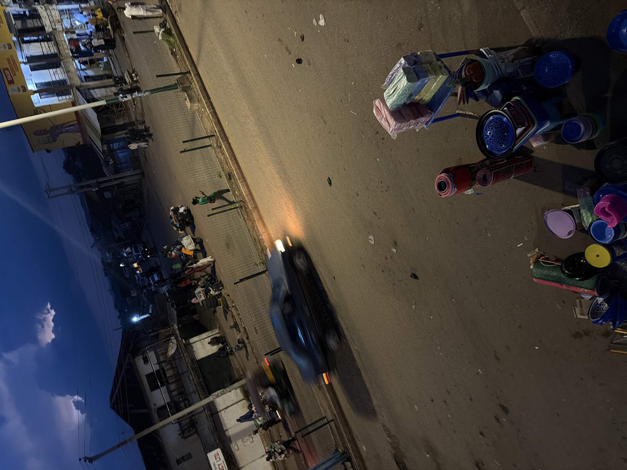 The image depicts a street scene at dusk in Bukuru, Nigeria. The sky is a deep blue with hints of light in the clouds, suggesting either twilight or an approaching storm. The street is paved and appears to be a busy thoroughfare.
In the foreground, a stack of colorful plastic items, including buckets and what appear to be mats or rolled-up textiles, are displayed on the side of the road. A blue cart with more items is also visible.
Further down the street, a dark-colored car is in motion, its headlights and taillights illuminated, with motion blur indicating speed. Other vehicles and a motorcycle with passengers are present in the midground. People are seen walking and gathered in small groups on the sidewalks and near the buildings.
The buildings lining the street are a mix of structures, some with multiple stories and businesses operating at street level, indicated by signage and open storefronts. A prominent yellow building with signage and a large mural is visible on the right side of the image. Power lines and poles crisscross the sky above the street. The overall impression is of a lively urban environment at the end of the day.