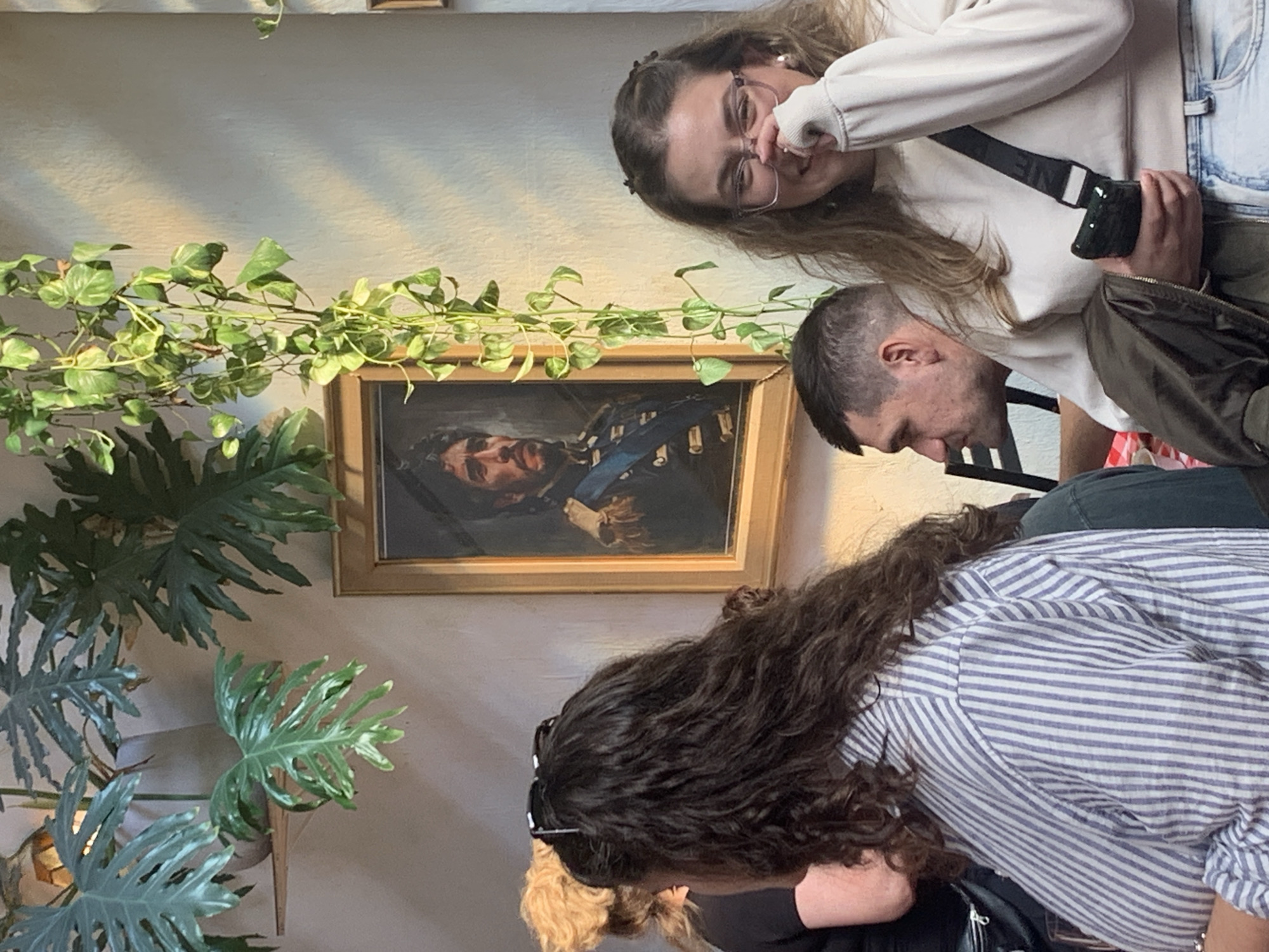 This indoor image features three individuals, a framed painting, and several plants, presented with a tilted perspective.
In the upper right, a young woman with long brown hair, wearing glasses and a light-colored sweatshirt over light-wash jeans, is looking towards the viewer. She is pinching her nose with one hand, possibly in amusement, while holding a dark-colored phone or object in the other. A black crossbody bag strap crosses her chest, with faint, partially obscured text that seems to read "NEW YORK."
Below her, a man with short, dark hair is shown in profile, looking downwards. He is wearing a dark shirt. In the foreground, partially visible, another person with long, dark, curly hair is seen from behind, bending forward. This individual is wearing a blue and white striped button-down shirt.
Central to the image is a framed portrait painting hanging on a light-colored wall. The painting depicts a man with dark hair and a beard, wearing a dark blue military-style jacket with lighter detailing.
The setting is adorned with greenery. A vine with small, variegated leaves trails across the wall above and to the left of the painting. To the left and below the painting, a large plant with deeply lobed, green leaves is prominent.
The lighting suggests a daytime setting. The overall atmosphere is casual, likely within a cafe or gallery, with the location context provided as Buenos Aires, Argentina.