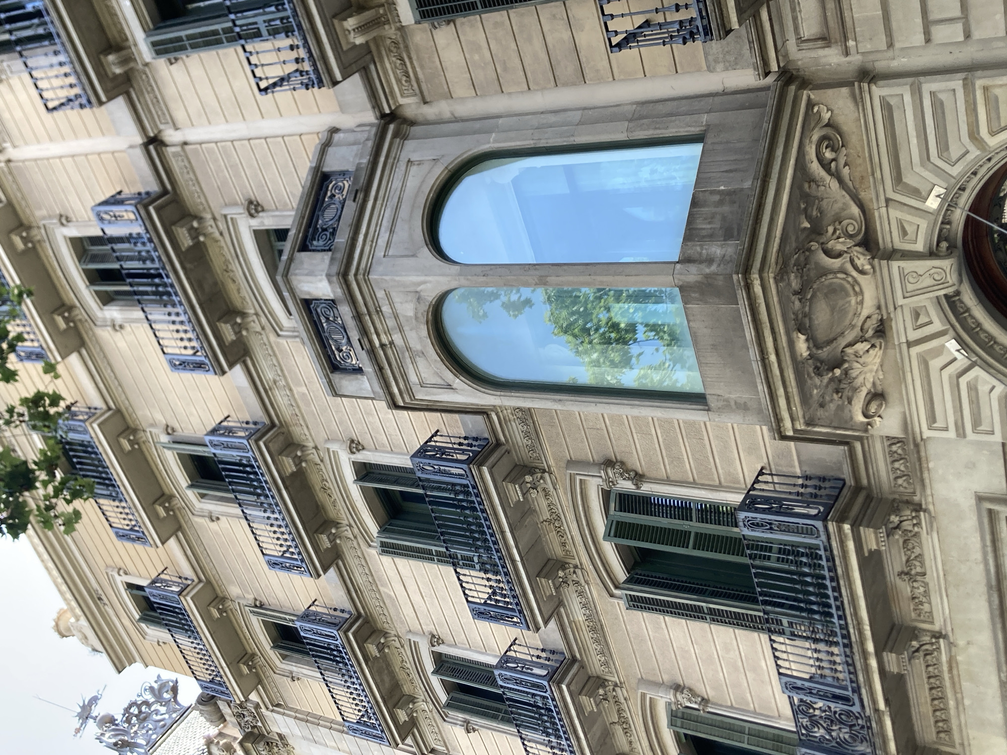 This image shows a close-up view of the facade of an ornate building, likely in Barcelona, Spain, given the architectural style. The building's exterior is constructed with light-colored stone, featuring decorative molding and intricate stone carvings. Multiple windows with dark green shutters are visible, some of which are partially open. Small balconies with dark blue wrought-iron railings project from some of the windows, adding to the building's detailed ornamentation.
Dominating the upper center of the image are two large, arched windows. The upper window reflects a clear blue sky, while the lower window reflects green foliage, suggesting the presence of trees nearby. The orientation of the image is such that the building appears to be viewed from a low angle, looking upwards.
The lighting suggests it is daytime, with natural light illuminating the building's facade. There are no people or animals visible in the frame, and the focus is solely on the architectural details of the building. The scene evokes a sense of historic grandeur and European urban architecture. No text is visible in the image.