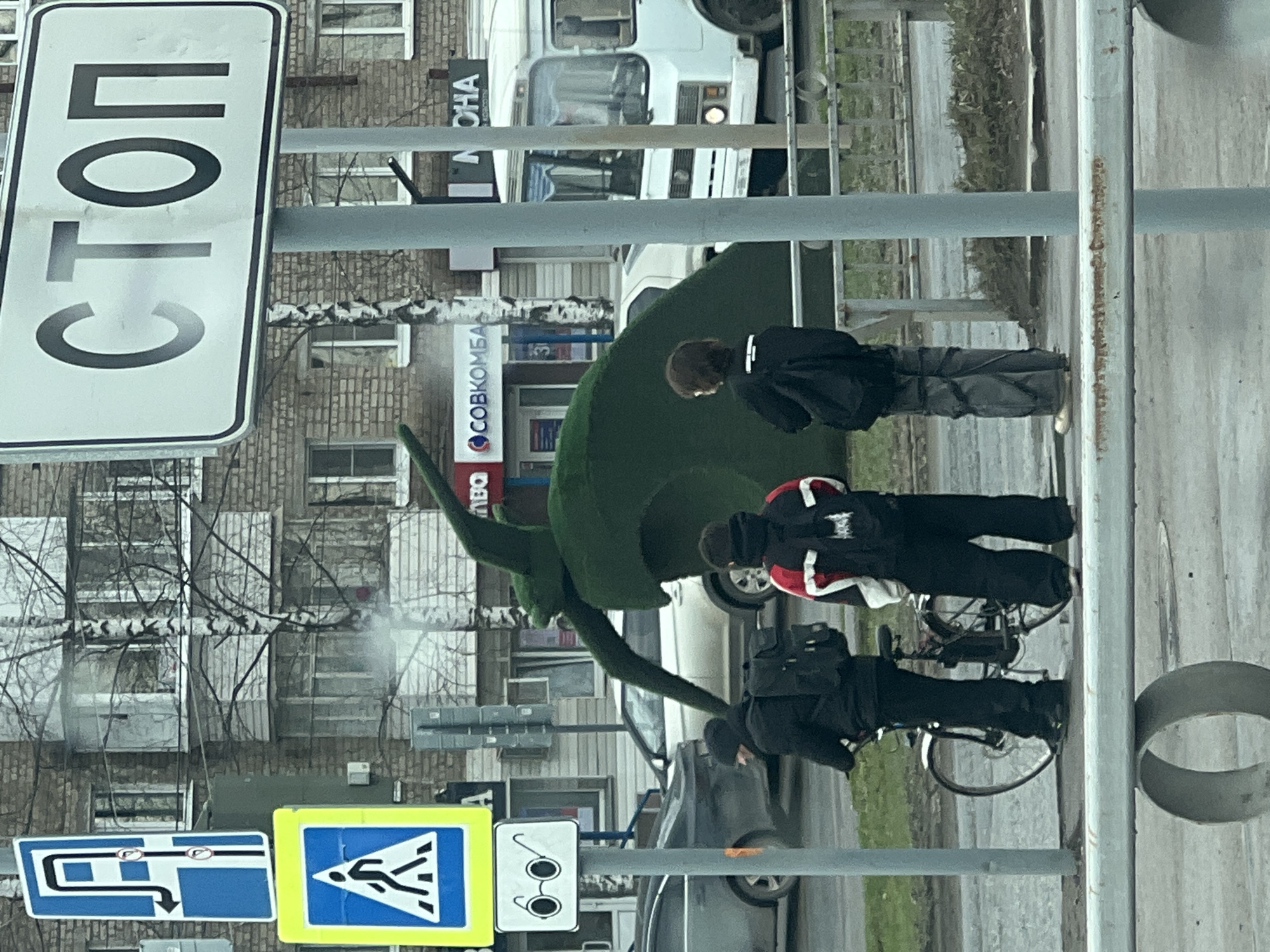 The image depicts a street scene in Chaikovsky, Russia. A large, green topiary sculpture of a dinosaur stands beside a sidewalk. Three individuals are present: one standing and two crouched near bicycles. The standing person is wearing dark clothing. The two crouched individuals also wear dark clothing, with one sporting a jacket with red and white accents. Traffic signs are visible, including a "СТОП" (STOP) sign and pedestrian crossing and parking signs. Buildings are present in the background, including one with signage for "СОВКОМБАНК" (SOVCOMBANK). A white van is parked on the street. The environment appears to be urban, with trees and pavement.