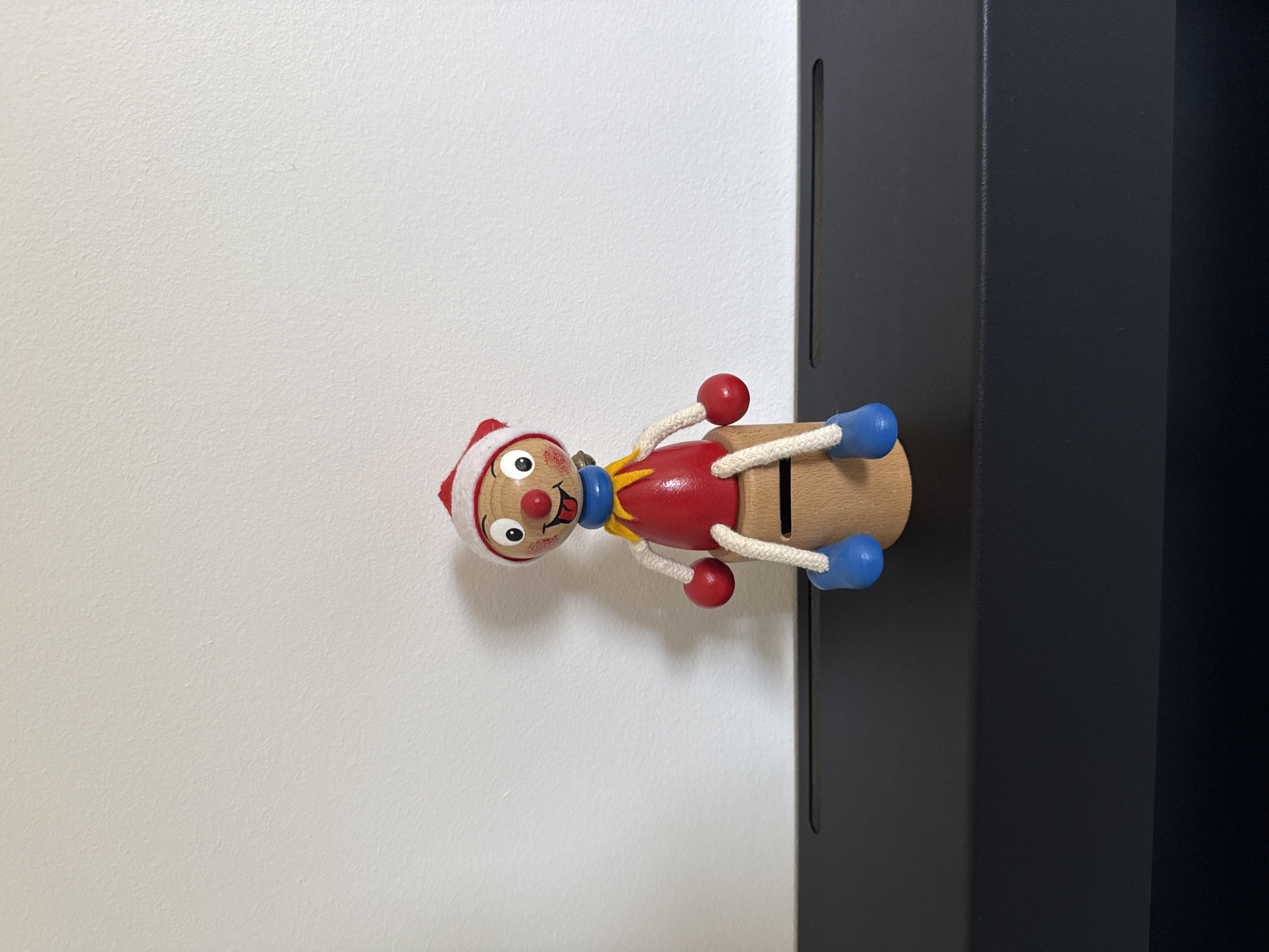 A wooden toy figure, resembling a clown with a red body, blue feet, and a red and white hat, sits on a black surface against a white wall. The figure has a painted face with a red nose, wide eyes, and a pink tongue sticking out. It is wearing a blue collar and a yellow ruff. The figure's arms are attached by white strings to red balls, and its legs are also made of blue balls attached by strings. The toy is positioned centrally in the frame, with the black surface extending horizontally across the bottom of the image. The white wall provides a plain background, with no visible features or decorations. The lighting appears to be natural, casting a soft shadow to the right of the toy. This scene is likely an indoor setting, potentially a home or a shop, and is not specific to any particular location within Drumettaz-Clarafond, France.