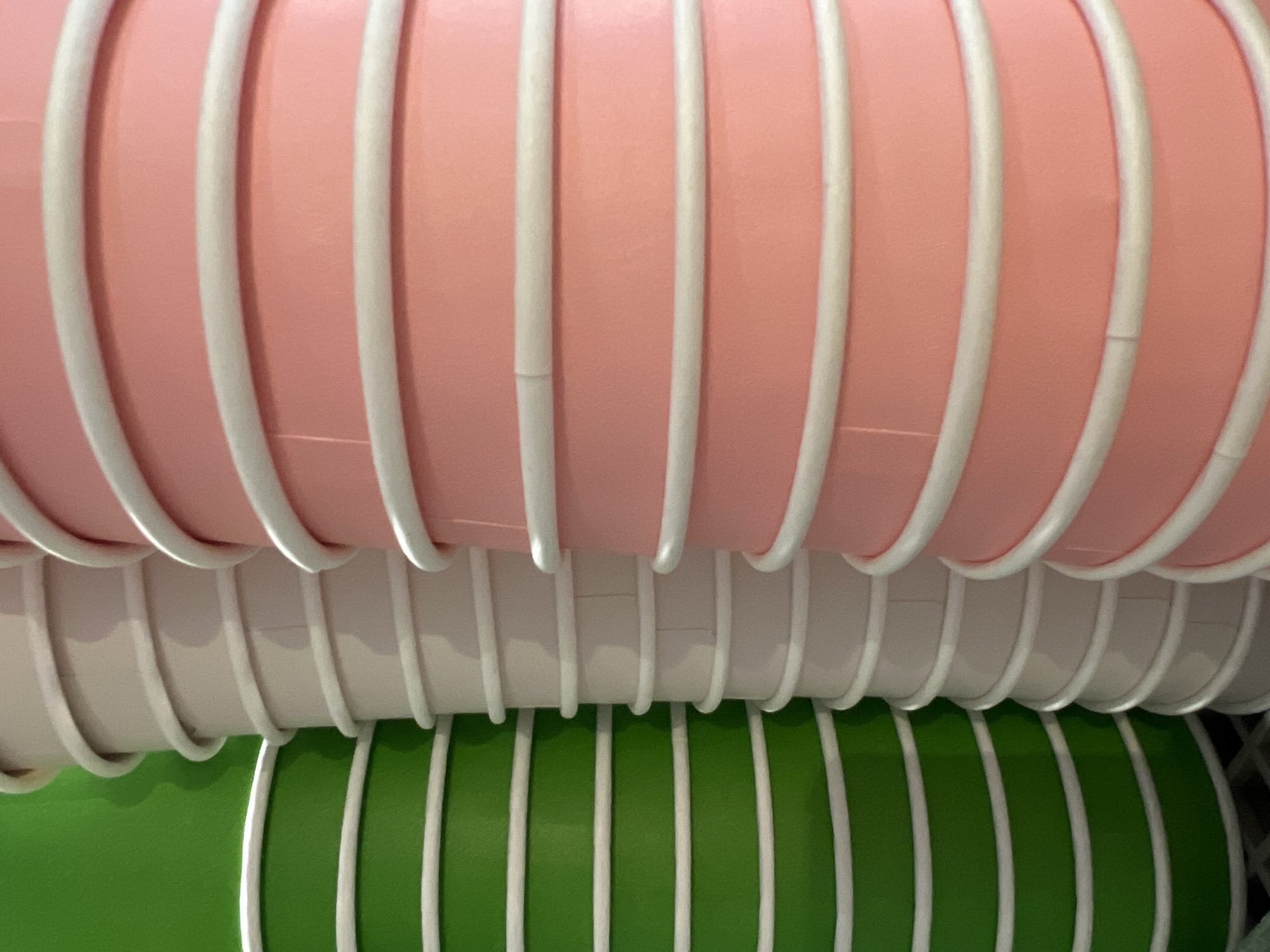 The image displays three stacks of disposable paper cups. The leftmost stack consists of green cups with white rims. The middle stack comprises white cups with white rims. The rightmost stack features pink cups with white rims. All cups appear to be identical in size and shape, and they are stacked vertically, filling the frame. The background is out of focus, with a hint of green on the left and a darker area behind the stacks. The lighting is even, highlighting the textures of the paper cups. No people or actions are depicted. The image is a close-up, focusing solely on the arrangement of these cups. It is not possible to determine the location within Moscow, Russia, from the visual information provided.