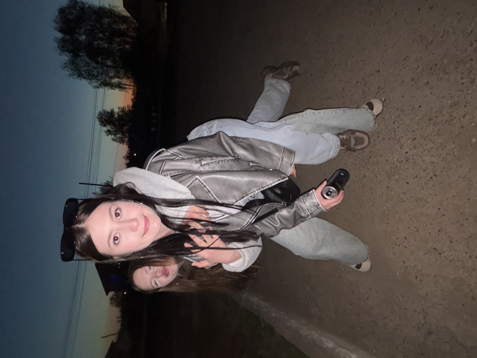 A low-angle, rotated photograph captures two women lying on a paved surface at dusk. The foreground woman, facing the camera, wears sunglasses atop her head, a grey hooded sweatshirt, and a metallic grey leather jacket. She holds a dark-colored can in her right hand. The second woman lies behind her, slightly lower in the frame, with a playful expression. Both are wearing light-colored jeans. They are lying on a dark asphalt road with sparse vegetation visible in the background. The sky exhibits hues of dusk, transitioning from orange near the horizon to dark blue above. The location, indicated as Фоки, Russia, is implied by contextual information and presumed given the request.