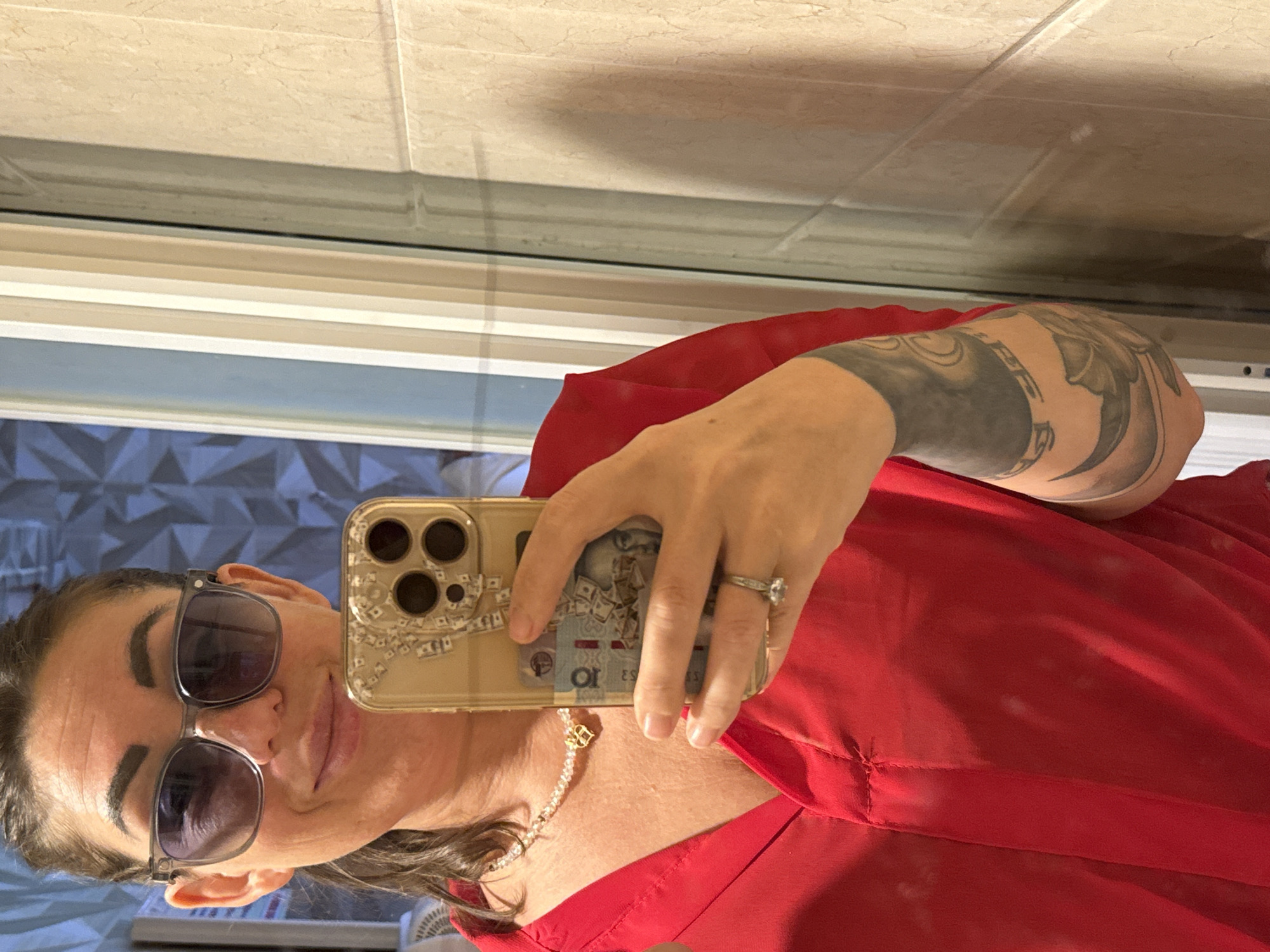 A woman in a red shirt takes a selfie in a bathroom. She is wearing large sunglasses and a necklace. Her phone is decorated with a money-themed case and she has a ring on her left hand. She has a tattoo on her left arm. The background shows tiled walls and a door frame. The location is not specified in the image or its context.