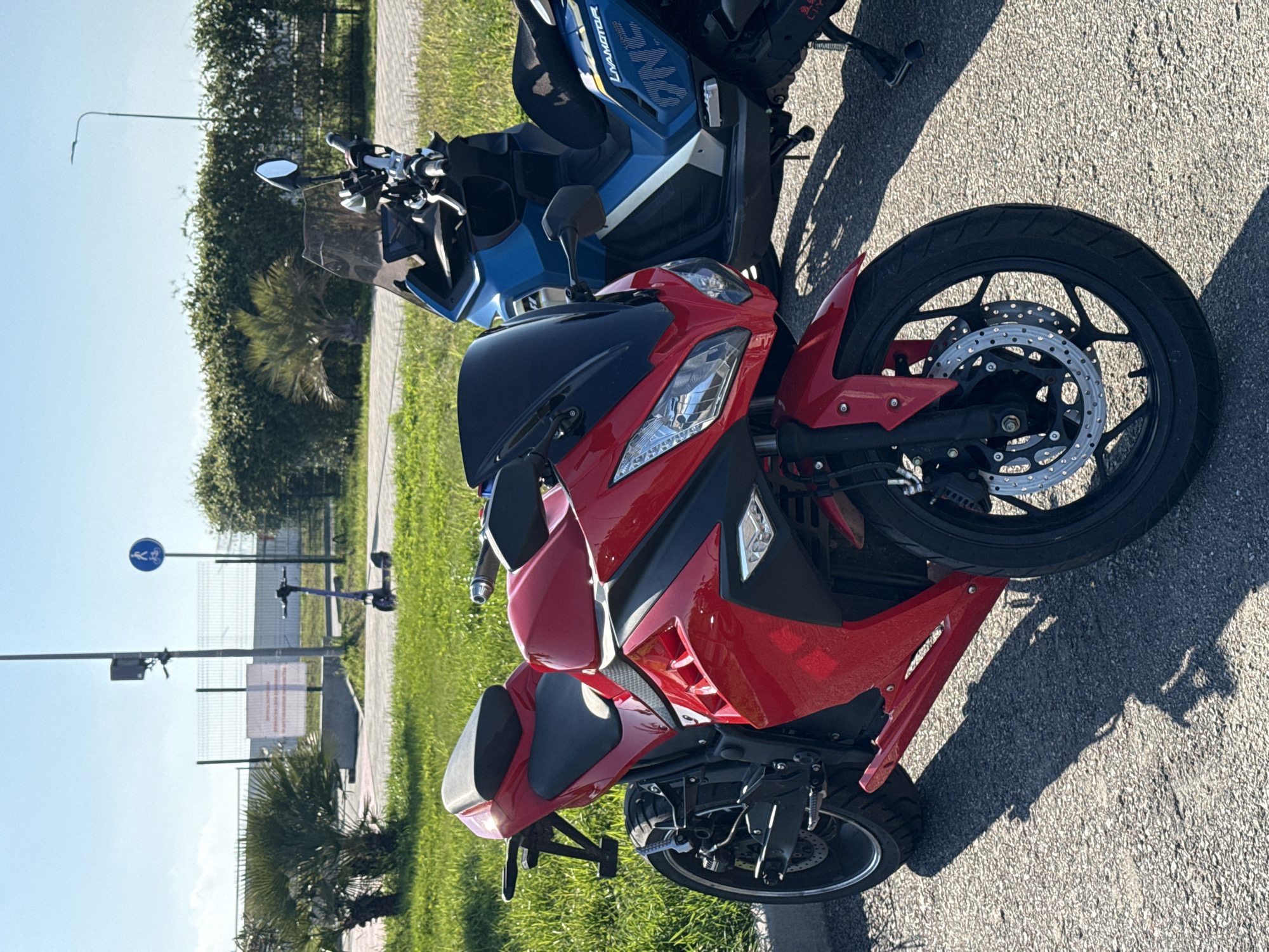The image captures two motorcycles parked outdoors on a paved surface in Sirius, Russia, under a bright, clear sky. In the foreground, a vibrant red and black sports motorcycle is prominently displayed, positioned facing right. Its front wheel, equipped with dual disc brakes, is clearly visible, along with its handlebars, windscreen, and sleek bodywork. Behind it, partially visible, is a blue and white scooter or larger motorcycle, bearing the text "UNAMOTOR" on its side panel.
The motorcycles are situated alongside a grassy area featuring dense green hedges and several palm trees, indicative of a warm climate. A paved sidewalk or pathway runs parallel to the grass, beyond which a metal fence separates the area from the background. A blue circular sign, displaying a white pictogram of a pedestrian and a bicycle, is mounted on a pole near the fence. Further back, a streetlight is visible, and a distant white sign with faint, unreadable red text can be seen through the fence.
The scene is bathed in bright sunlight, casting distinct shadows on the pavement, suggesting it is daytime, likely morning or late afternoon. There are no people visible, and the focus is on the parked vehicles and their immediate surroundings.