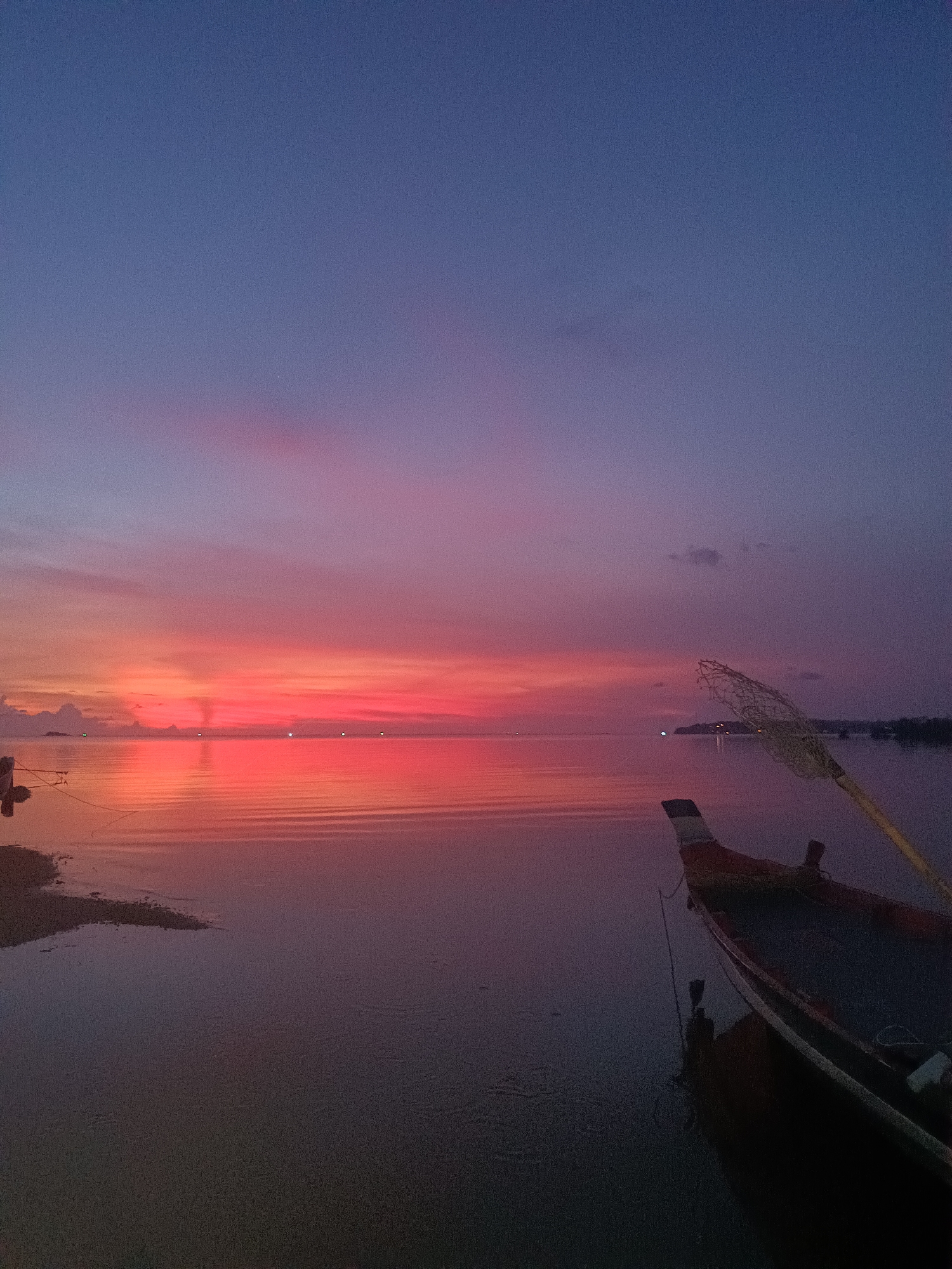 The image captures a tranquil scene at dusk in Baan Wok Tum, Thailand. The sky exhibits a gradient of colors, transitioning from a deep blue at the top to shades of pink and red near the horizon. This colorful reflection is visible on the calm water's surface. A small, partially visible landmass is on the left, while a boat is anchored in the water near the right foreground. A fishing net extends from the boat. The overall environment suggests a serene, coastal setting as the day transitions to night.