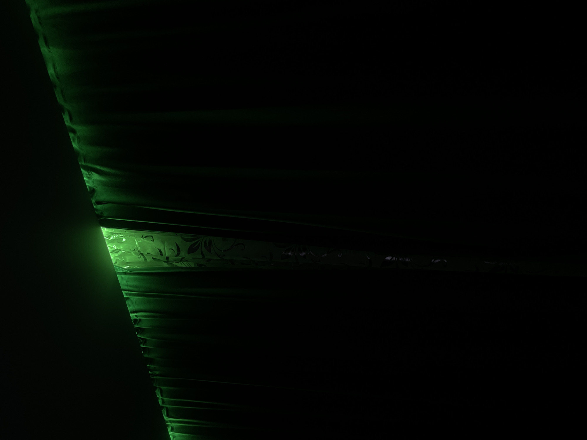 The image shows a portion of an object illuminated by green light. The object has a textured surface with horizontal indentations, and in one section, there is a design with floral elements. The overall scene is dark, with the green light being the primary source of illumination. The environment is unspecified and could be indoors. There is no information about the location in the city of Chaikovsky, Russia, people, or any actions or interactions.