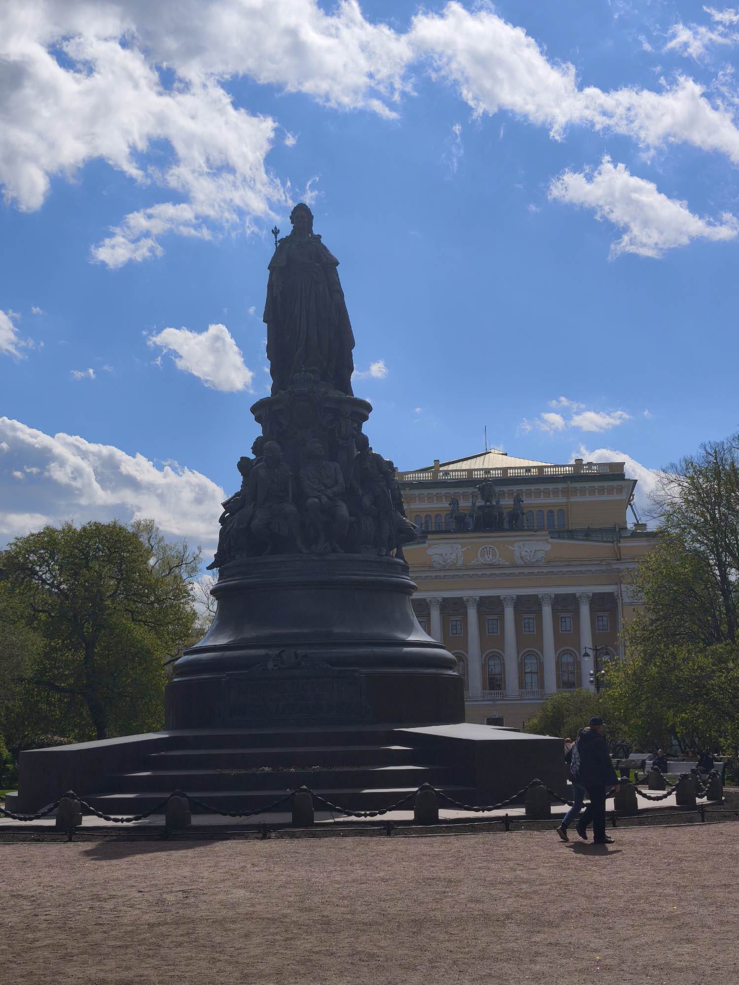 The image depicts a sunny outdoor scene in Saint Petersburg, Russia. At the center is the Monument to Catherine the Great, a large, dark bronze sculpture of the Empress, standing atop a multi-tiered pedestal adorned with figures. The monument is encircled by a low fence made of posts connected by heavy chains.
Behind the monument is a yellow and white classical building with ornate details, including columns and statues on its roof. Trees with green leaves frame the building on either side.
In the foreground, the ground is covered with a reddish-brown gravel. Two individuals are walking across the gravel in front of the monument.
The sky is blue with fluffy white clouds. The light indicates it is likely daytime.