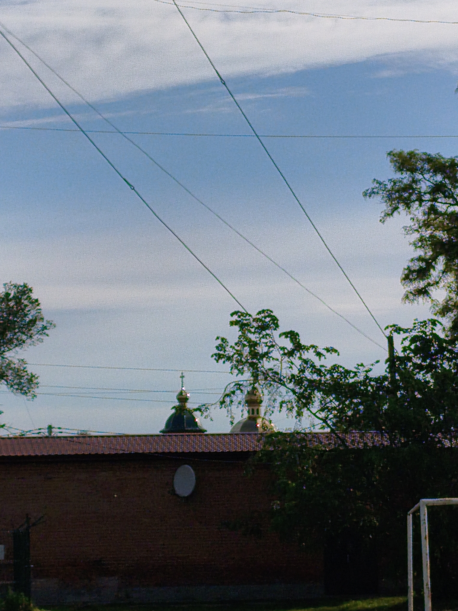 The image presents a daytime outdoor scene, likely in a semi-rural or provincial setting in Ustyvytsia, Ukraine. The dominant feature of the upper portion is a light blue sky with scattered, wispy white clouds. Crisscrossing the sky are several lines, composed of what appear to be individual, discrete dots, running diagonally from the top left to the bottom right, and one horizontally. These lines might represent cables, wires, or even a netting structure.
In the middle ground, a long, dark red brick wall with a reddish-brown corrugated roof stretches across the frame. Behind this wall and above its roofline, two golden onion domes, characteristic of Eastern Orthodox churches, are visible. The dome on the left is mostly obscured, while the one further right is more prominent and features a cross atop its spire, confirming its religious purpose. Green foliage from trees partially frames these domes and extends from the left and right sides of the image.
Mounted on the red brick wall, towards the right side, is a small, white, circular object, which appears to be a satellite dish. At the very bottom right corner of the image, partially hidden by trees, the white metal frame of a soccer goal is visible. A patch of green grass can be seen at the bottom of the frame, suggesting a field or yard.
The lighting indicates it is daytime, under a partly cloudy sky. There are no people visible, and no specific activity or event is depicted. No discernible text is present in the image.