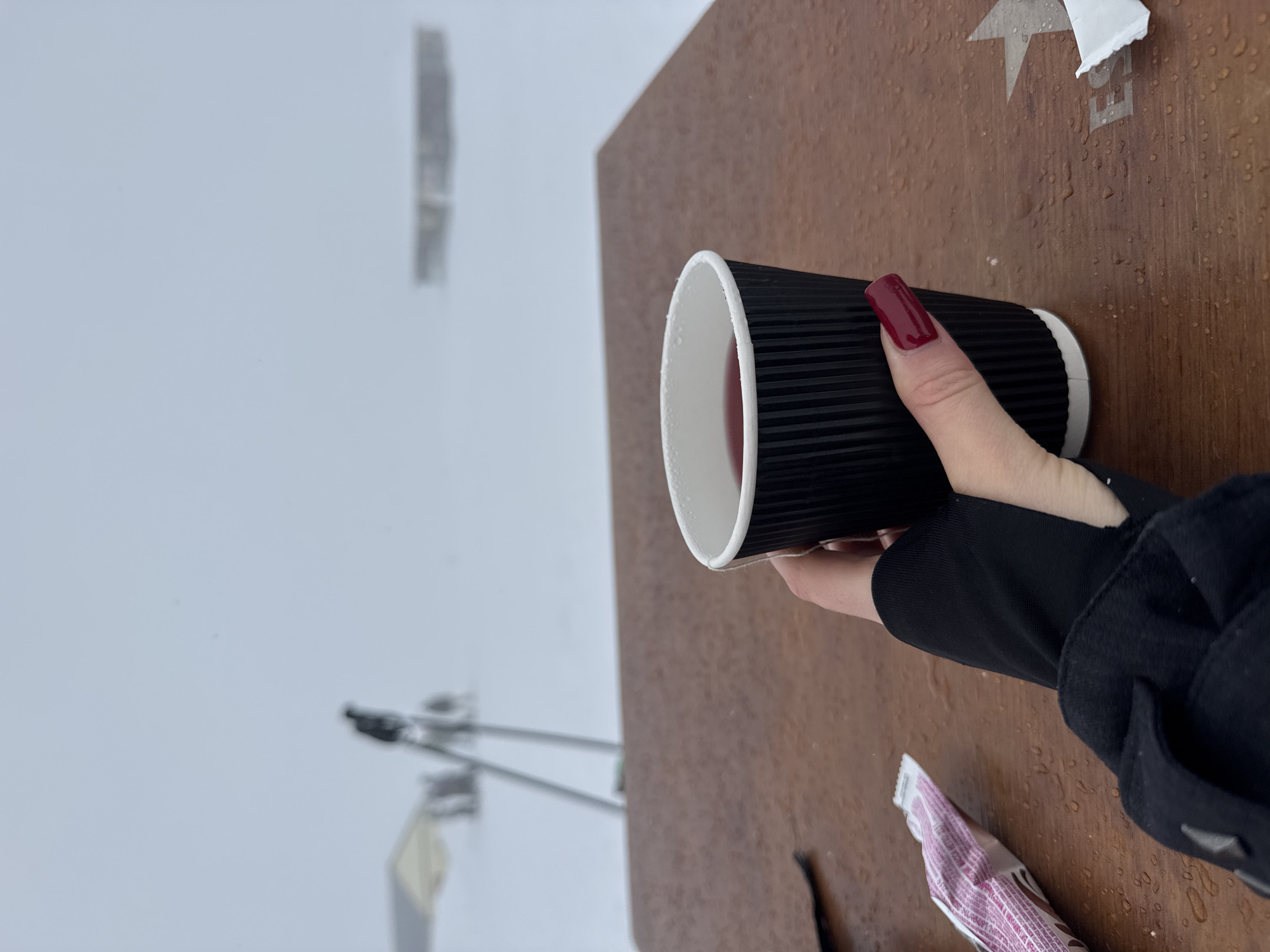 A person holding a black corrugated paper cup is captured in this medium shot. The background features an overcast or foggy sky, possibly indicating a snowy or cold environment. In the distance, blurred architectural structures and lampposts are visible, suggesting a city location. The cup is filled with a red liquid, possibly coffee, and held by a hand with a burgundy nail. The person is wearing a black jacket. The cup and hand are positioned over a brown surface, which seems to be an outdoor table, with raindrops and some paper wrappings scattered on it. The specific city of Unknown, Unknown, is not discernible from the given visual information.