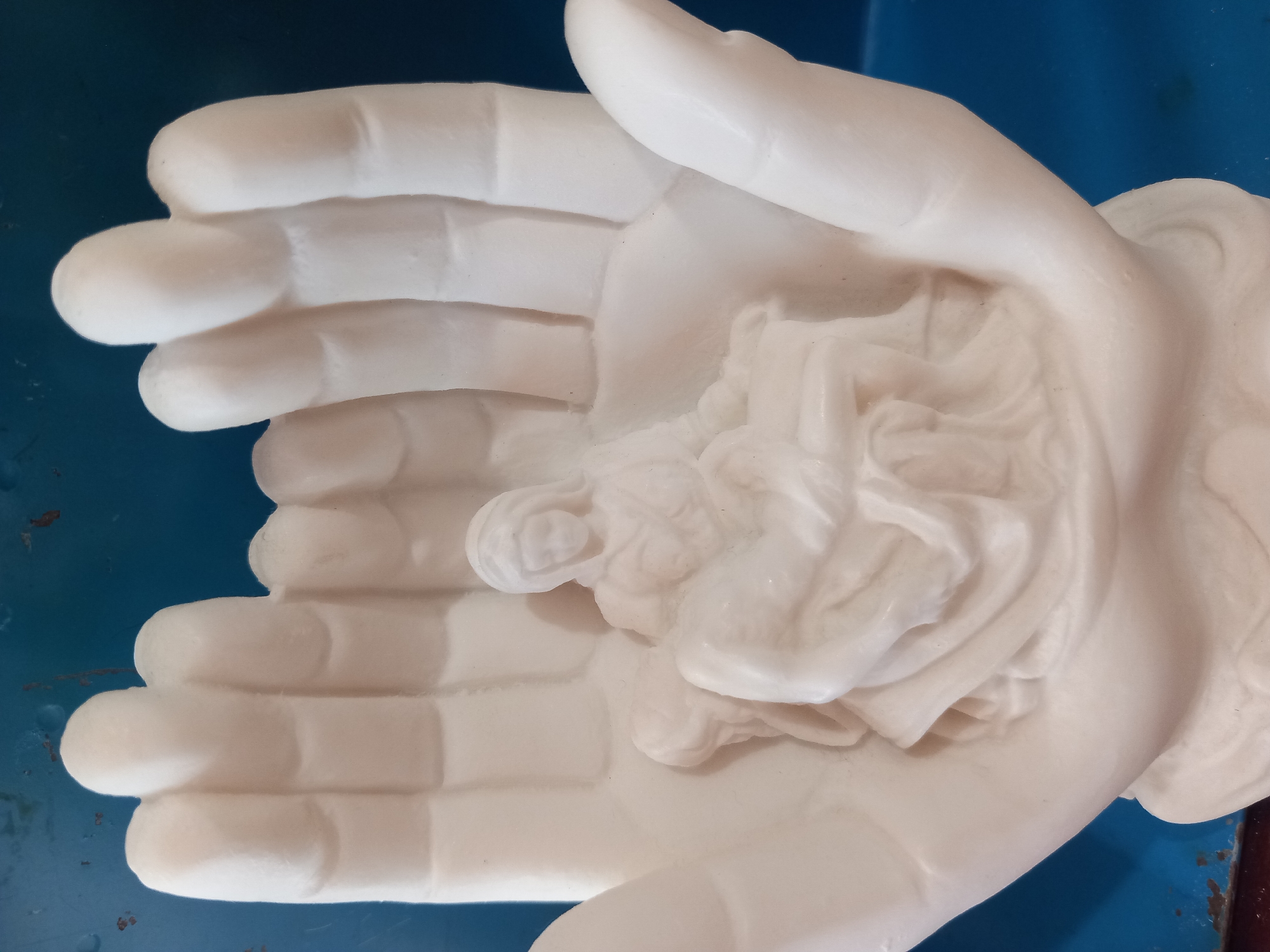A close-up, eye-level, static shot shows a white, marble-like sculpture of a pair of cupped hands holding a Pietà. The sculpture is centered in the frame. The hands are open, with fingers pointing upwards, and appear to be supporting the figure within. The Pietà depicts a woman, presumably the Virgin Mary, holding the body of Jesus. The woman is seated and draped in a flowing gown, with her head bowed. The body of Jesus is cradled in her lap, appearing lifeless. The sculpture is well-detailed, with folds in the fabric and delicate facial features. The background is a solid, dark blue. The lighting is soft and even, highlighting the texture and form of the sculpture. The image does not contain any identifiable location information within a city.