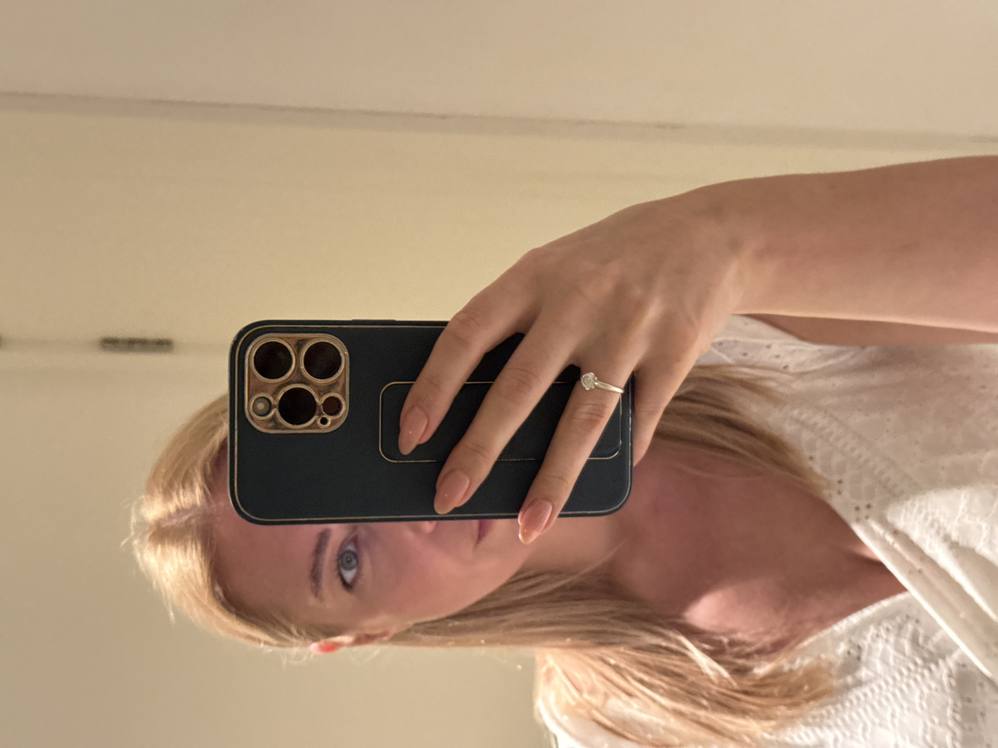 A blond woman is taking a selfie in front of a mirror. She is holding a dark-colored smartphone with a gold-colored camera frame in her right hand, partially covering her face. Her left hand is visible, adorned with a ring. She is wearing a white top. The background is a neutral-colored wall. There is no definitive information in the image to indicate this photo was taken in Ghadeer Al Tayr, United Arab Emirates.