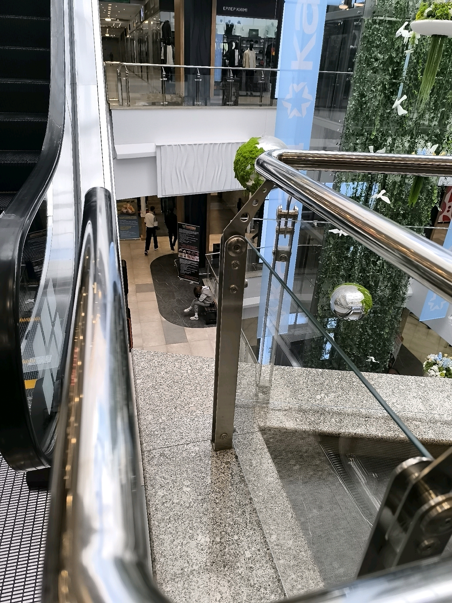 An interior view of a modern shopping mall shows an escalator on the left and a glass railing on the right, leading down. In the background, people are visible walking and sitting. A large green wall with artificial plants covers a section of the mall, with white bird figurines attached. A reflective sphere hangs near the green wall. The flooring is a speckled granite. Signs and store displays are visible in the upper levels of the mall.