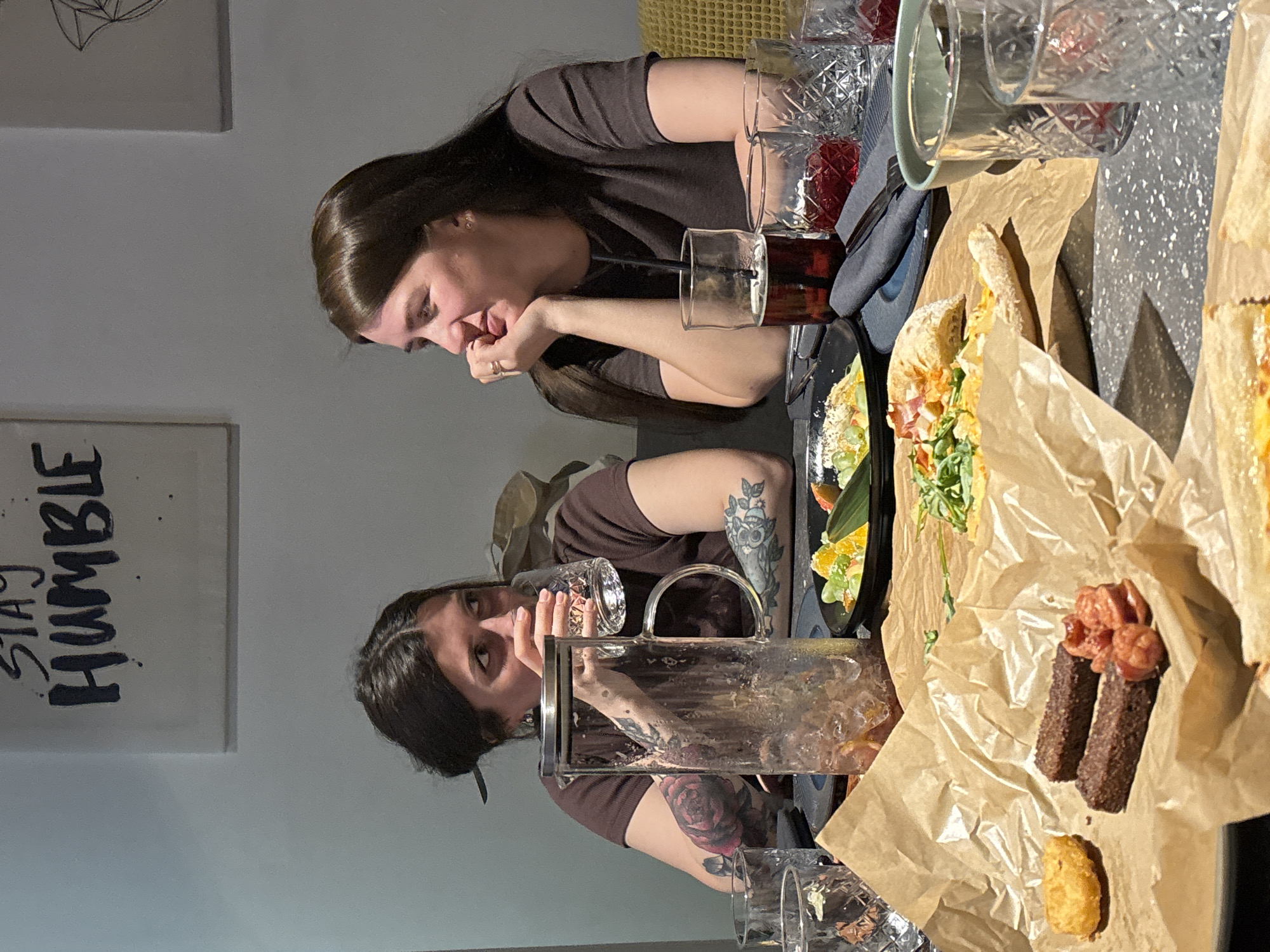 The image captures an indoor dining scene, likely in a restaurant or cafe in Chaikovsky, Russia, characterized by warm artificial lighting and a casual atmosphere. The central subjects are two women, one in the foreground and her reflection mirroring her presence on a dark, glossy table surface.
The primary woman, with long dark hair and wearing a dark brown short-sleeved top, rests her chin on her hand, looking downwards with a contemplative expression. A ring is visible on her left hand. Her reflection, seen from a different angle, shows her holding a patterned glass, appearing to look up and to the side. This reflected image prominently displays tattoos on her arms: one on her upper arm resembles an owl or floral design, while another on her forearm depicts a large red rose.
The table is laden with food and beverages. A glass containing a dark red drink with a straw is positioned near the foreground woman. Nearby are several other glasses, some empty, and a large glass pitcher filled with ice, suggesting a shared drink. The food items are largely wrapped in brown paper, indicative of a casual presentation style. Visible dishes include what appears to be a submarine sandwich or wrap filled with greens and other ingredients, as well as distinct portions of fried dark bread sticks topped with reddish pieces (possibly sausages or bacon), and a single golden-brown fried item. A small black plate with a garnish or salad is also visible.
In the background, a light-colored wall features two framed pictures. The lower frame clearly displays the stylized text "STAY HUMBLE" in a dark font. The upper frame has a geometric, perhaps abstract, design. The overall impression is that of friends or companions enjoying a meal and drinks in a relaxed setting.
