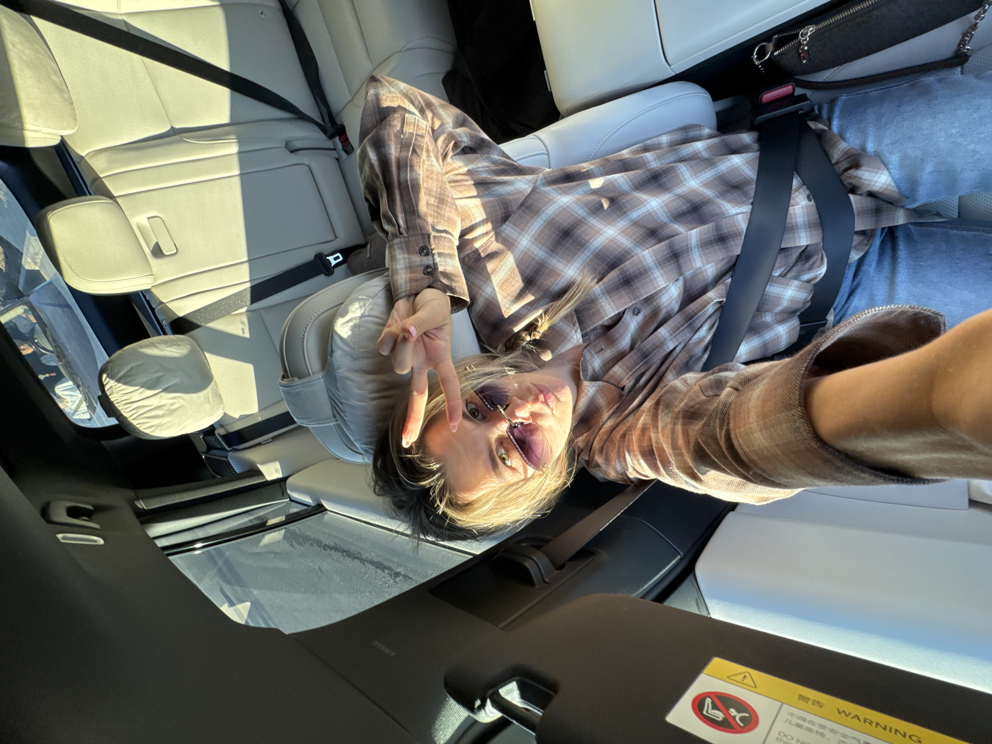 A young woman with blonde hair is lying on the back seat of a car, taking a selfie. She is wearing a brown and white plaid shirt and blue jeans. She has on sunglasses with purple lenses and is making a peace sign with her right hand. Sunlight is streaming into the car, casting shadows on the cream-colored leather seats. The car appears to be parked, possibly in a sunny outdoor location. The scene is casual and relaxed. A warning sticker is visible on a dark surface in the foreground, containing an icon of a child in a car seat and text in English and Chinese. The English text reads "WARNING DO NOT". The time of day is likely daytime due to the bright sunlight.